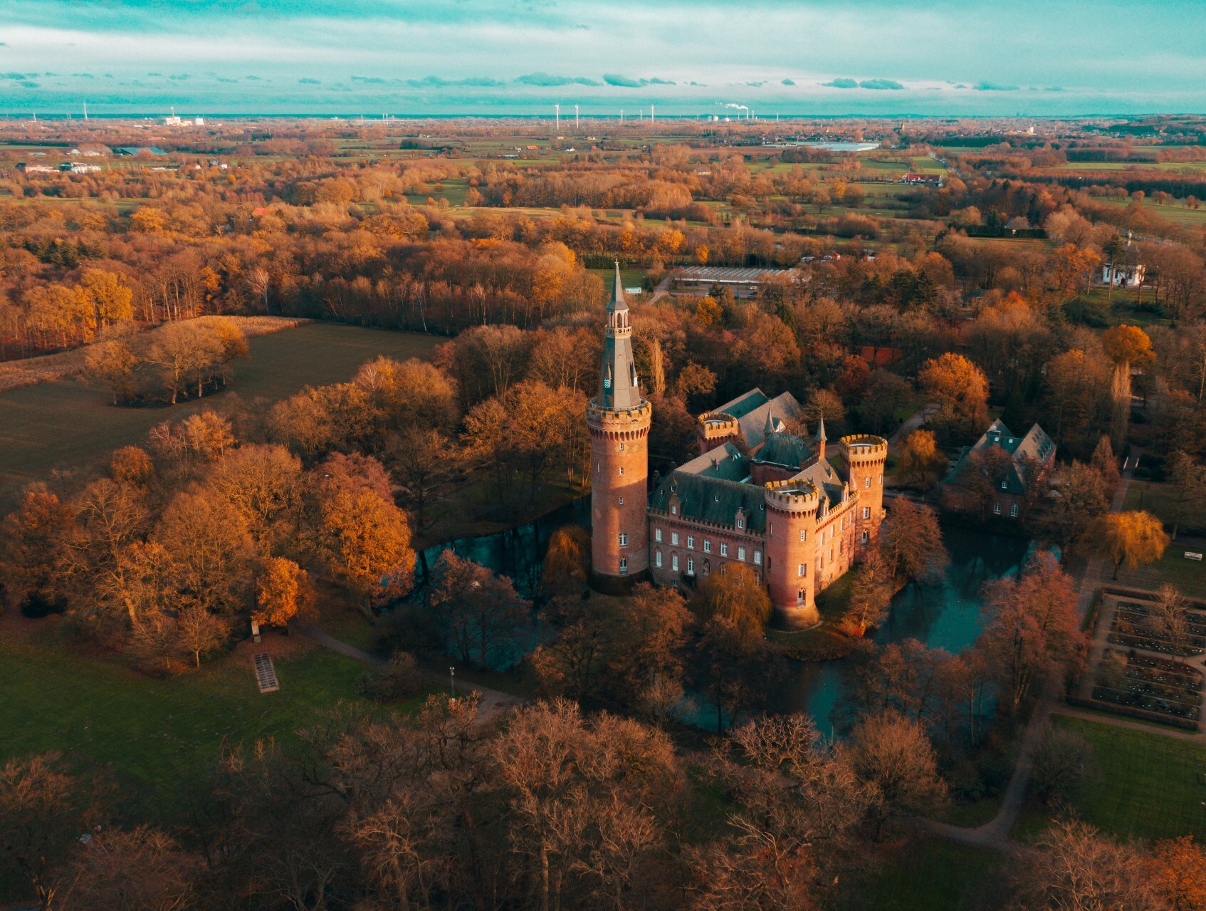 Drohnenaufnahme von Schloss Moyland in Bedburg-Hau am Niederrhein, umgeben von in rotes Licht getauchten Wäldern und Feldern