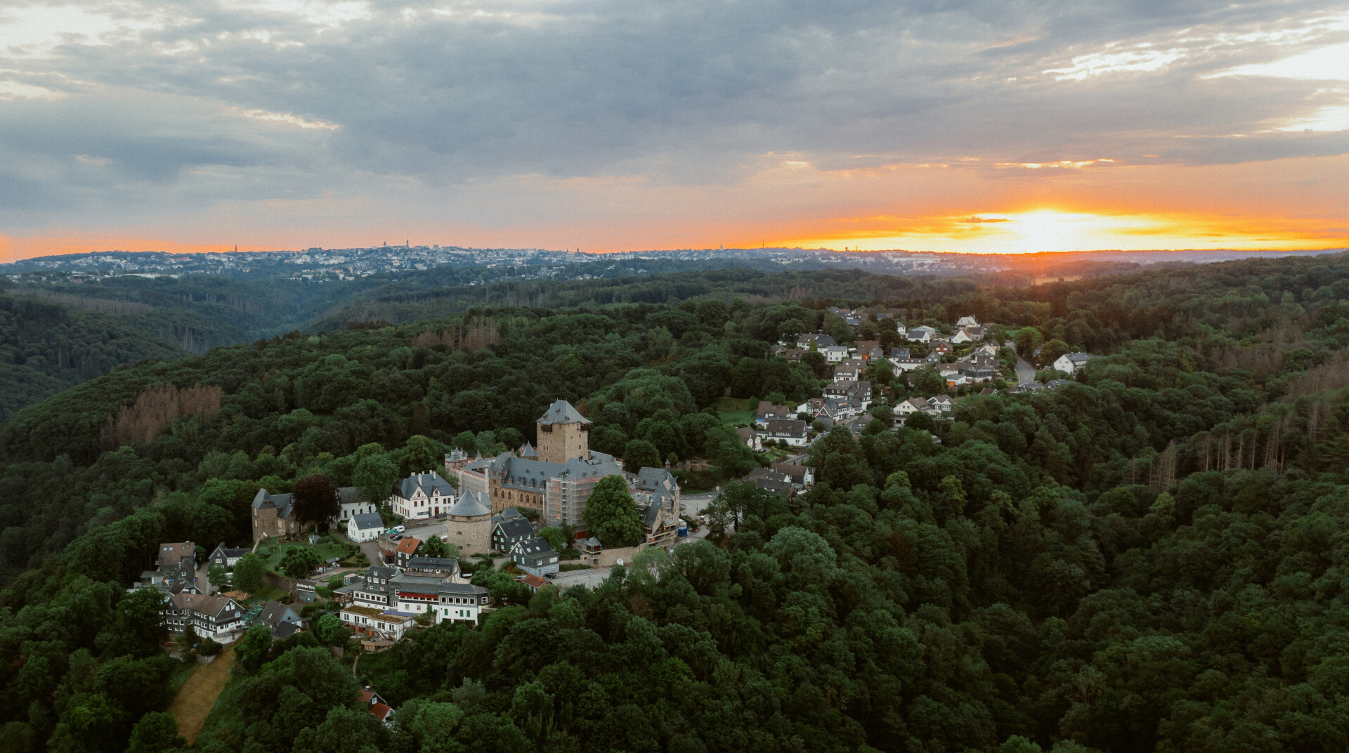 Schloss Burg umgeben von einzelnen Häusern und Wald aus der Vogelperspektive im Sonnenuntergang.