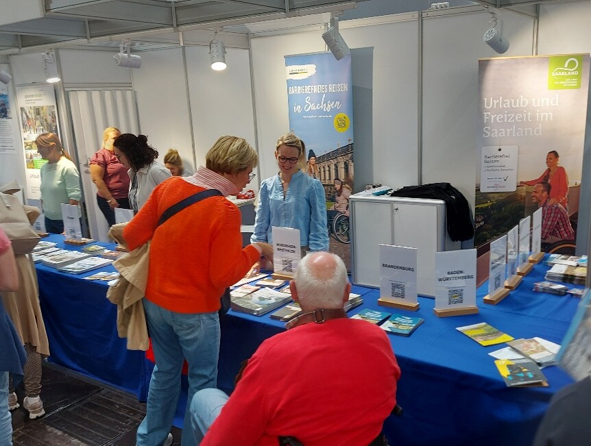 Eine Mitarbeiterin des Tourismus NRW steht auf der Rehacare in Düsseldorf an einem Tisch, auf dem viele Prospekte über das Reiseland Nordrhein-Westfalen liegen.  Vor dem Tisch sind eine Frau und ein Mann im Rollstuhl, die auf die Prospekte schauen.  