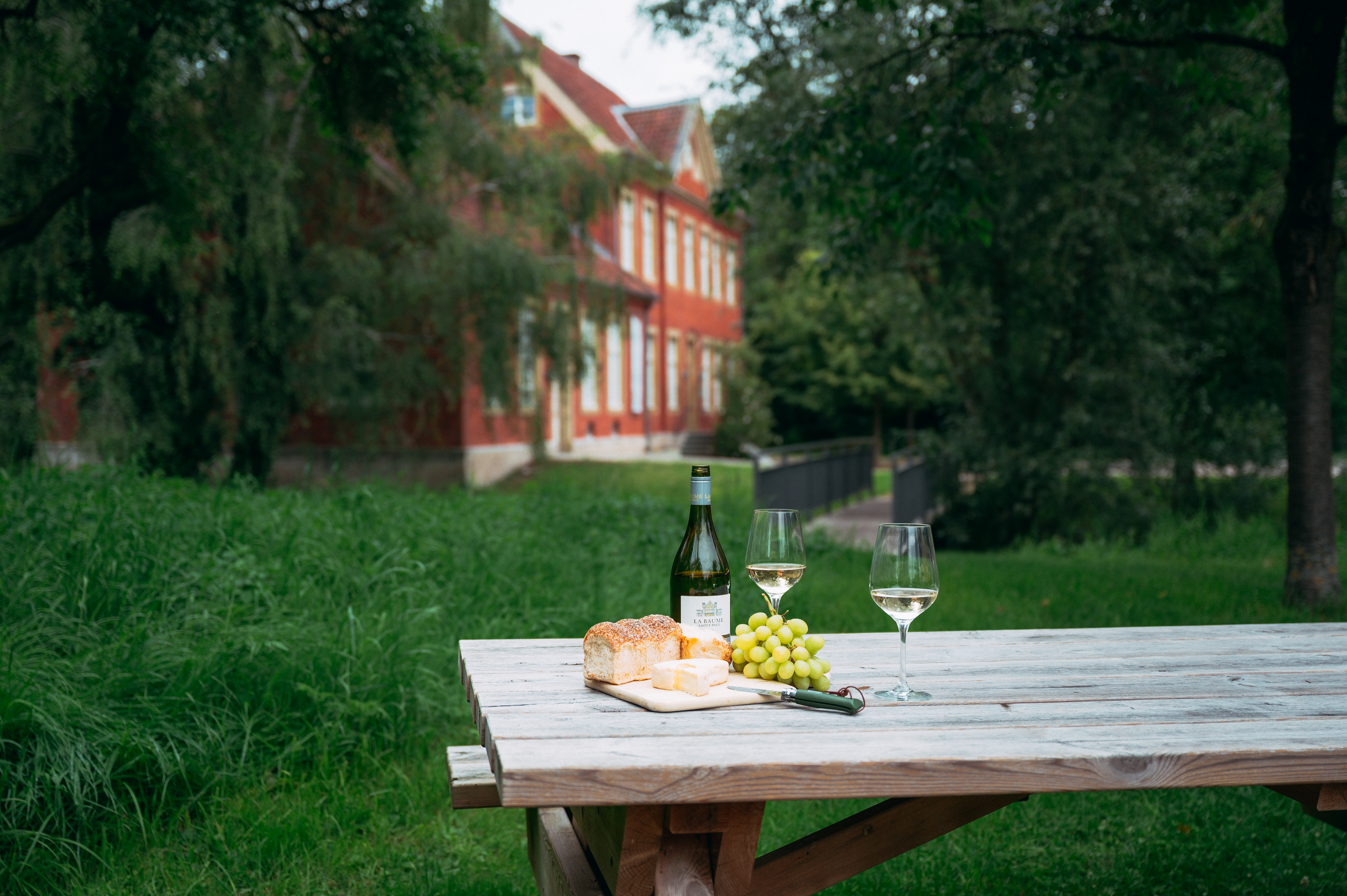 Im Park von Kulturgut Haus Nottbeck stehen eine Flasche Wein, zwei Weingläser, Trauben und Brot auf einem Holztisch. Das rote Haus ist im Hintergrund zu sehen. 