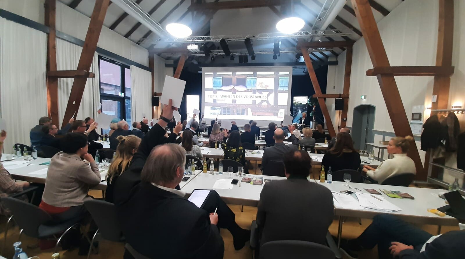 Menschen sitzen an Tischen und heben ihre Stimmzettel bei der Wahl des neuen Vorstands des Tourismus NRW auf der Mitgliederversammlung des Verbands 2022 in die Luft.