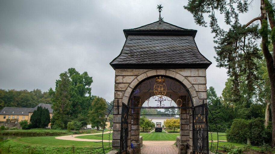 Ein steinernes Torhaus steht im Park von Schloss Dyck in Jüchen am Niederrhein auf einer steinernen Brücke. Im Hintergrund ist die gepflegte Gartenanlage von Schloss Dyck sichtbar. Auf beiden Seiten eines Weges und der Brücke ist viel Grün zu sehen, darunter Bäume und Rasenflächen. 