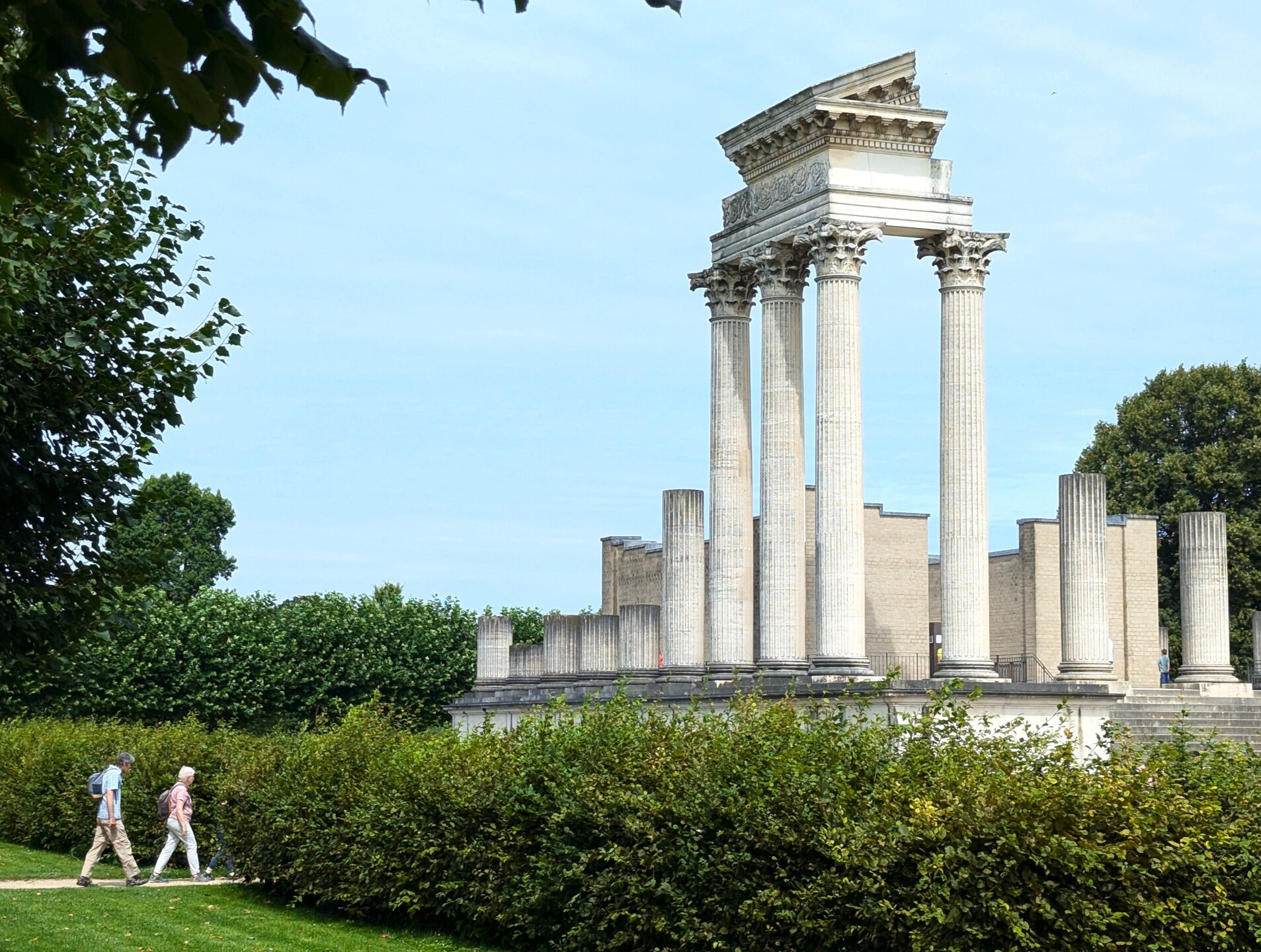 Hafentempel im Archäologischen Park Xanten © Tourismus NRW e.V./Maximilian Hulisz Zwei Menschen gehen auf einem Schotterweg an einer Hecke entlang, im Hintergrund ist die rekonstruierte römische Hafentempelanlage im Archäologischen Park Xanten am Niederrhein zu sehen. Die ruinenhafte Anlage besteht aus mehreren Säulen mit einem Giebel sowie weiteren Gebäudeteilen aus hellem Stein. Die Szene ist von Bäumen und gepflegtem Rasen umgeben.
