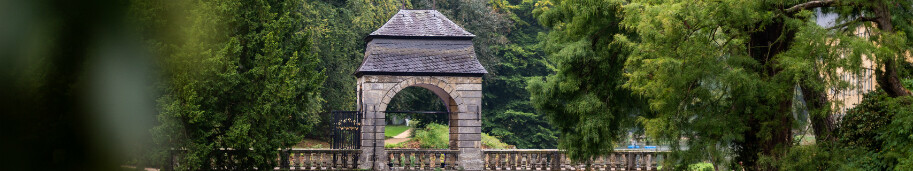 Ein steinernes Torhaus steht im Park von Schloss Dyck in Jüchen am Niederrhein auf einer Brücke, die ein ruhiges Gewässer überquert. Die Brücke hat mehrere Bögen, die sich im Wasser spiegeln. Das Torhaus ist von dichtem Grün umgeben, darunter große Bäume und Büsche. 
