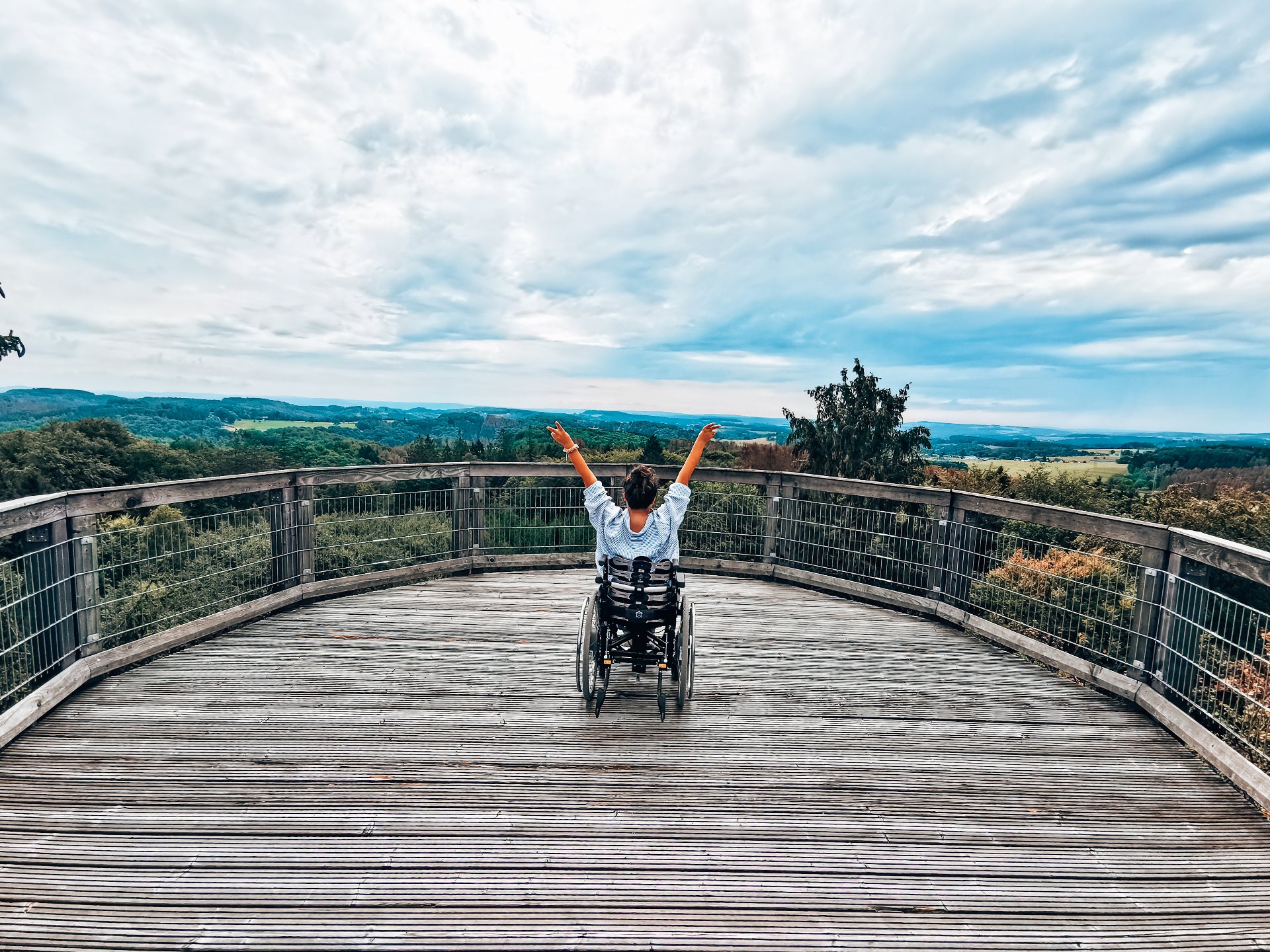 Eine Rollstuhlfahrerin schaut von der Aussichtsplattform des Baumwipfelpfads Panabora auf die grünen Hügel im Bergischen Land und recht die Arme in die Höhe.  