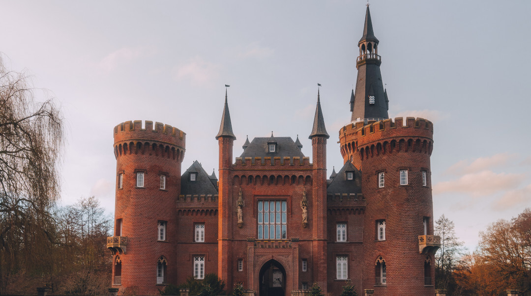 Backsteinbau Schloss Moyland am Niederrhein mit runden Ecktürmen, spitz zulaufenden Türmchen und einem hohen Turm. Davor liegt ein gepflegter Garten mit kugelförmig geschnittenen Büschen und Laub auf einem Rasen.
