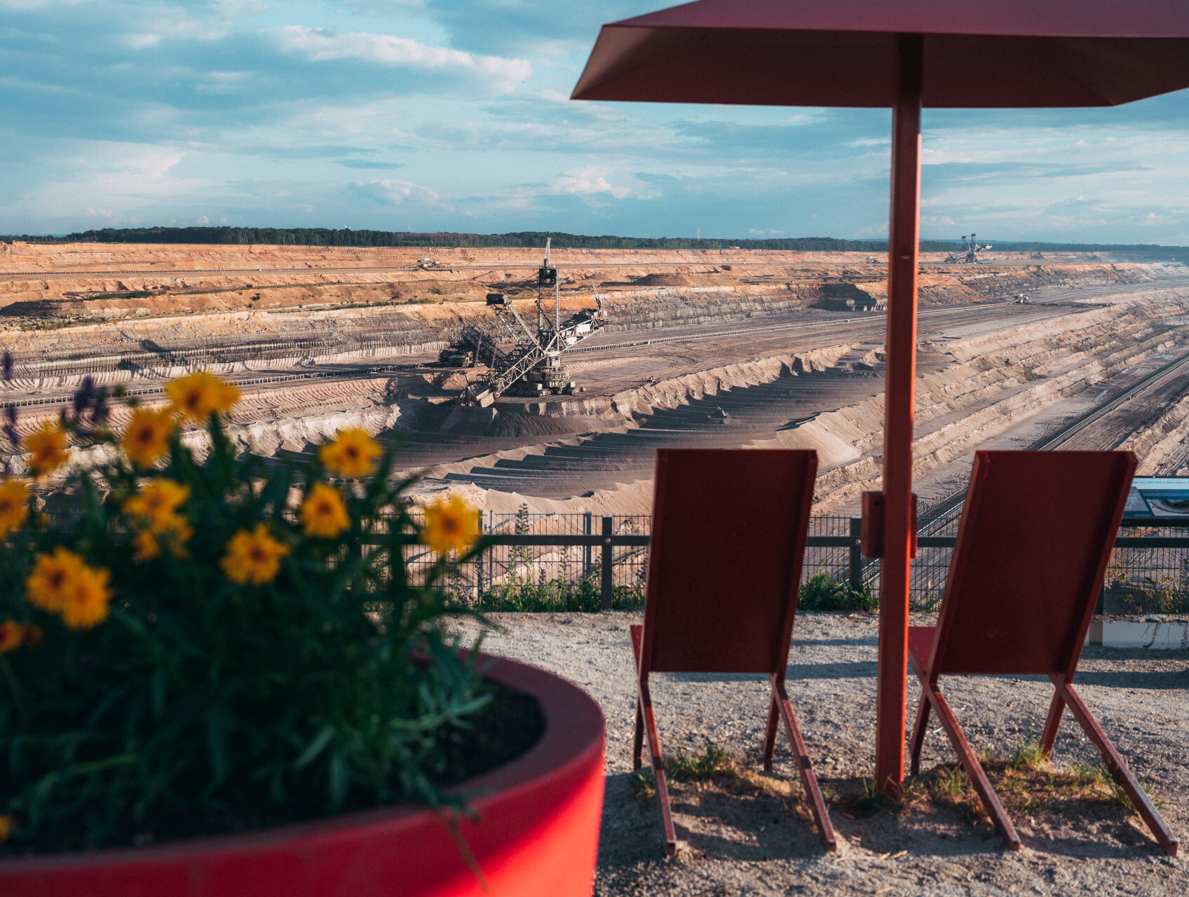 Zwei rote Liegestühle unter einem Sonnenschirm mit Blick auf den Braunkohletagebau Garzweiler im Rheinischen Revier; im Vordergrund ein großer Blumenkübel mit gelb und violett blühenden Blumen, im Hintergrund ein riesiger Schaufelradbagger in der gestuften Landschaft.