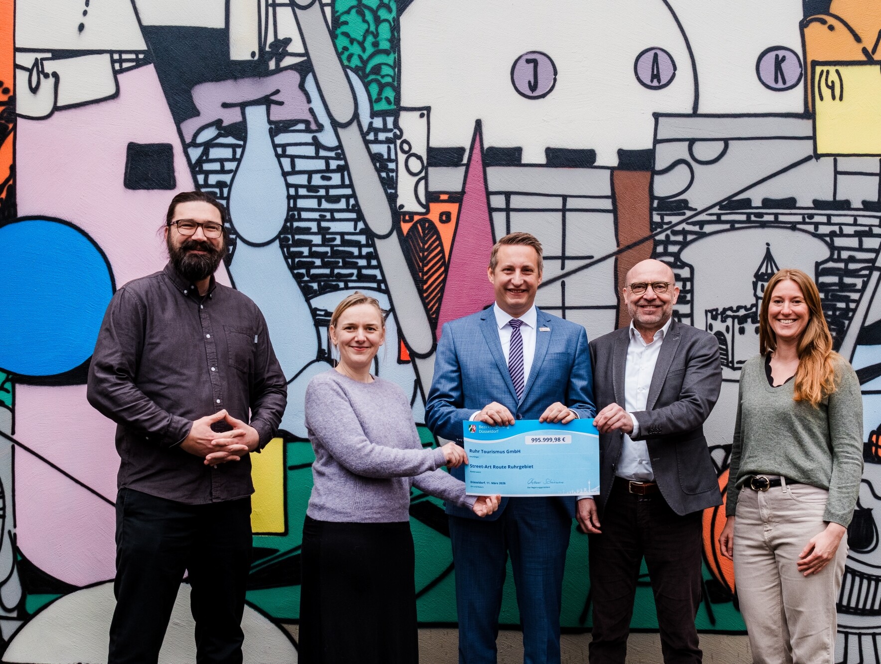 Fünf Menschen stehen vor einem großflächigen Street-Art-Werk. Drei von ihnen halten gemeinsam einen Förderbescheid in den Händen. 