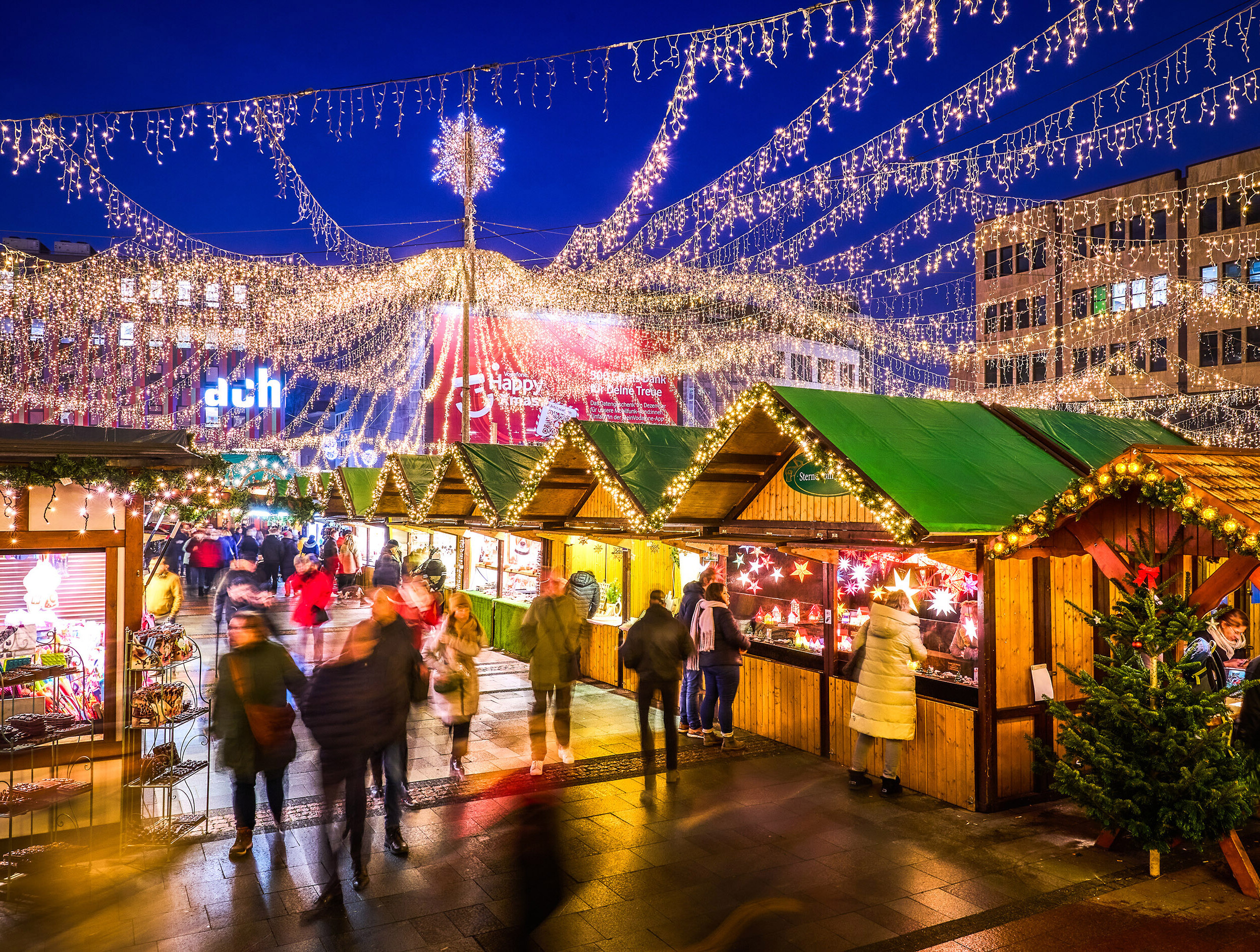 Festlich beleuchteter Internationaler Weihnachtsmarkt in Essen am Abend. Über den Holzhütten sind Lichterketten gespannt, die sich strahlenförmig von einem zentralen Punkt mit einem leuchtenden Stern aus nach außen verteilen und den Markt in ein warmes, gemütliches Licht tauchen. Besucher schlendern durch die Gänge zwischen den Buden, die verschiedene Produkte anbieten. 