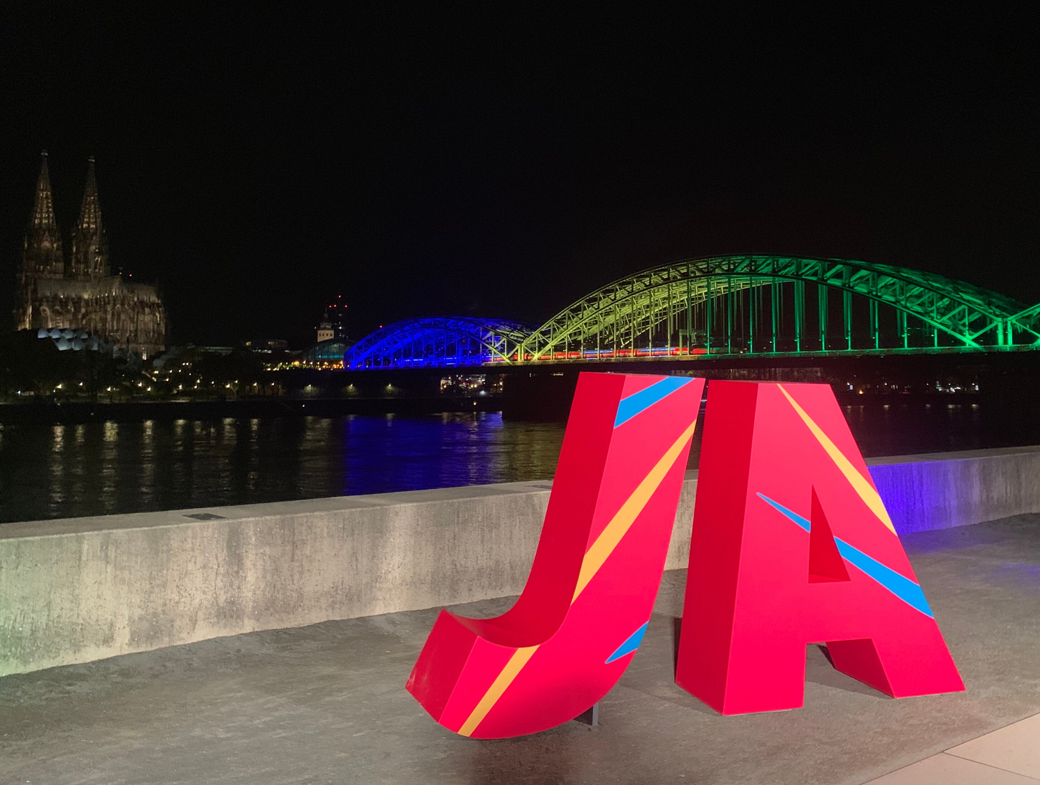 Vor der in den Farben der olympischen Ringe beleuchteten Hohenzollernbrücke und dem Dom in Köln steht ein großes rotes "Ja".