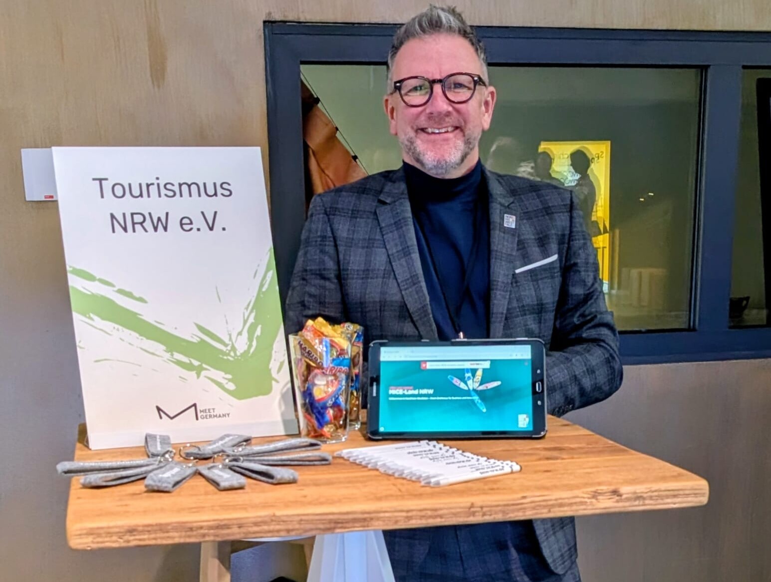 Ein Mitarbeiter von Tourismus NRW steht beim Meet Germany Summit NRW 2025 an einem Hochtisch. Darauf steht ein Aufsteller mit der Aufschrift "Tourismus NRW e.V." sowie ein Tablet. 