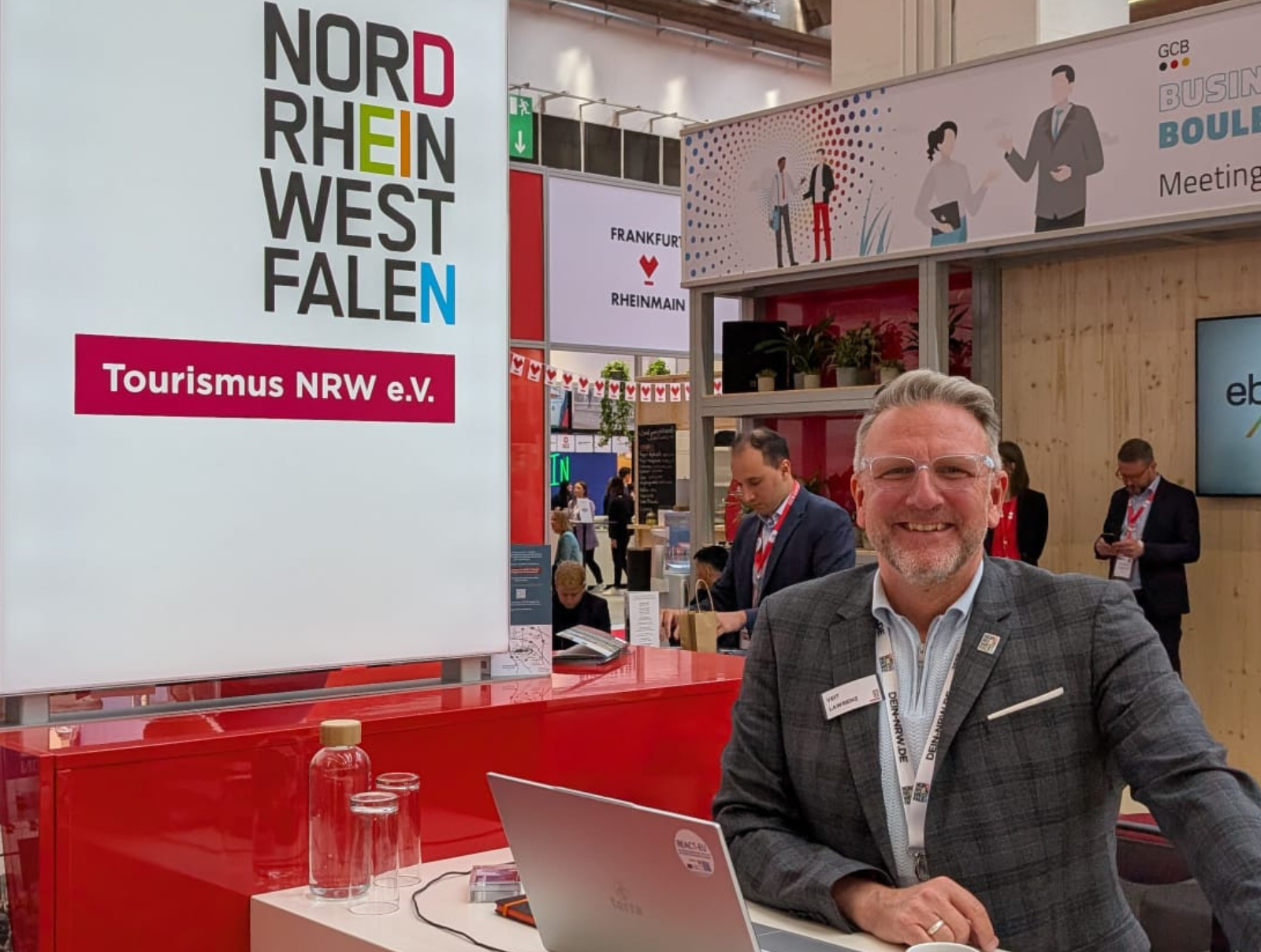 Ein Mitarbeiter von Tourismus NRW steht an einem Counter auf der IMEX in Frankfurt. Auf einer Wand neben ihm ist das Logo des Verbands. 