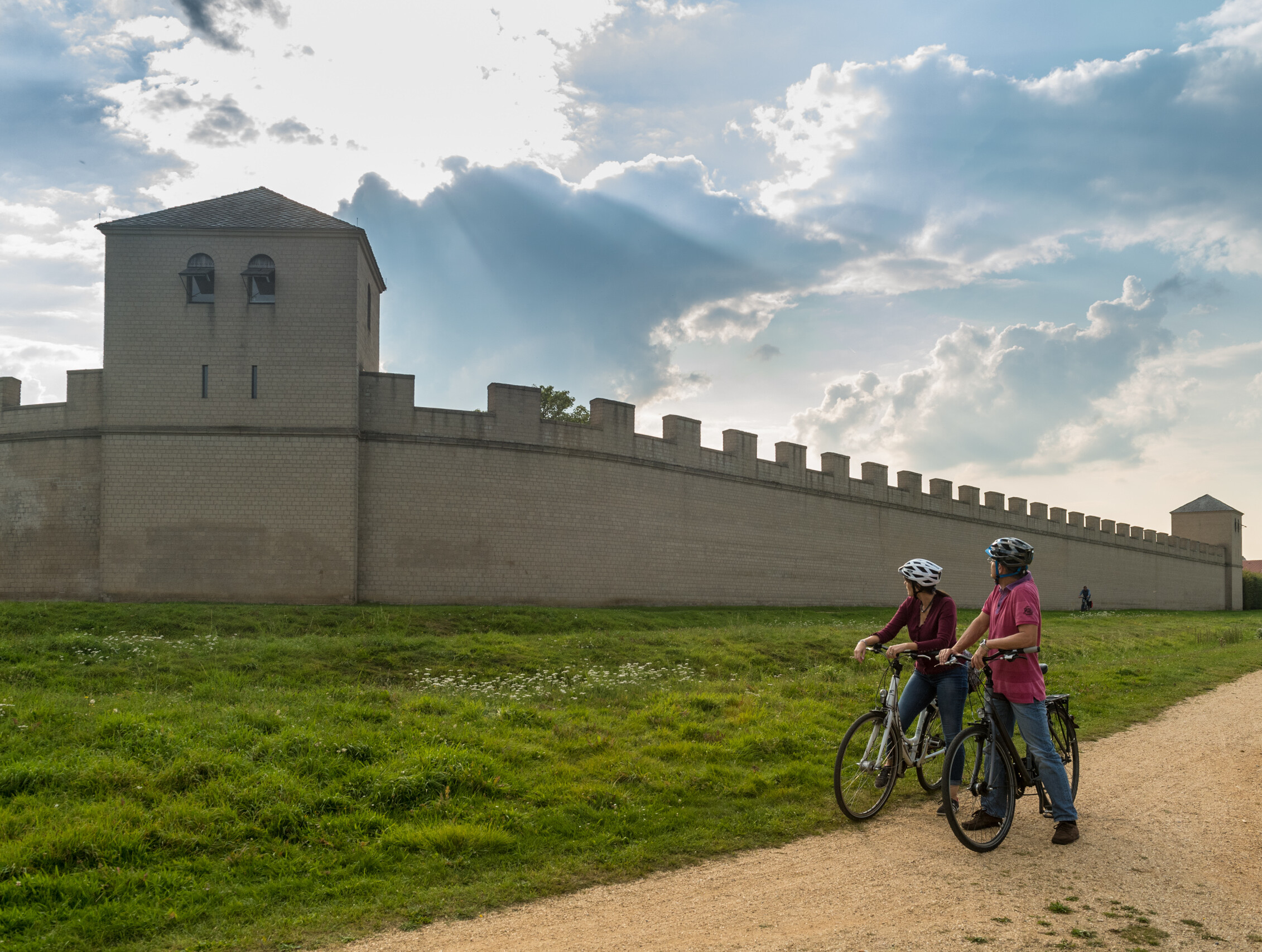 Zwei Menschen mit Fahrrädern schauen auf die Stadtmauer im Archäologischen Park Xanten. 