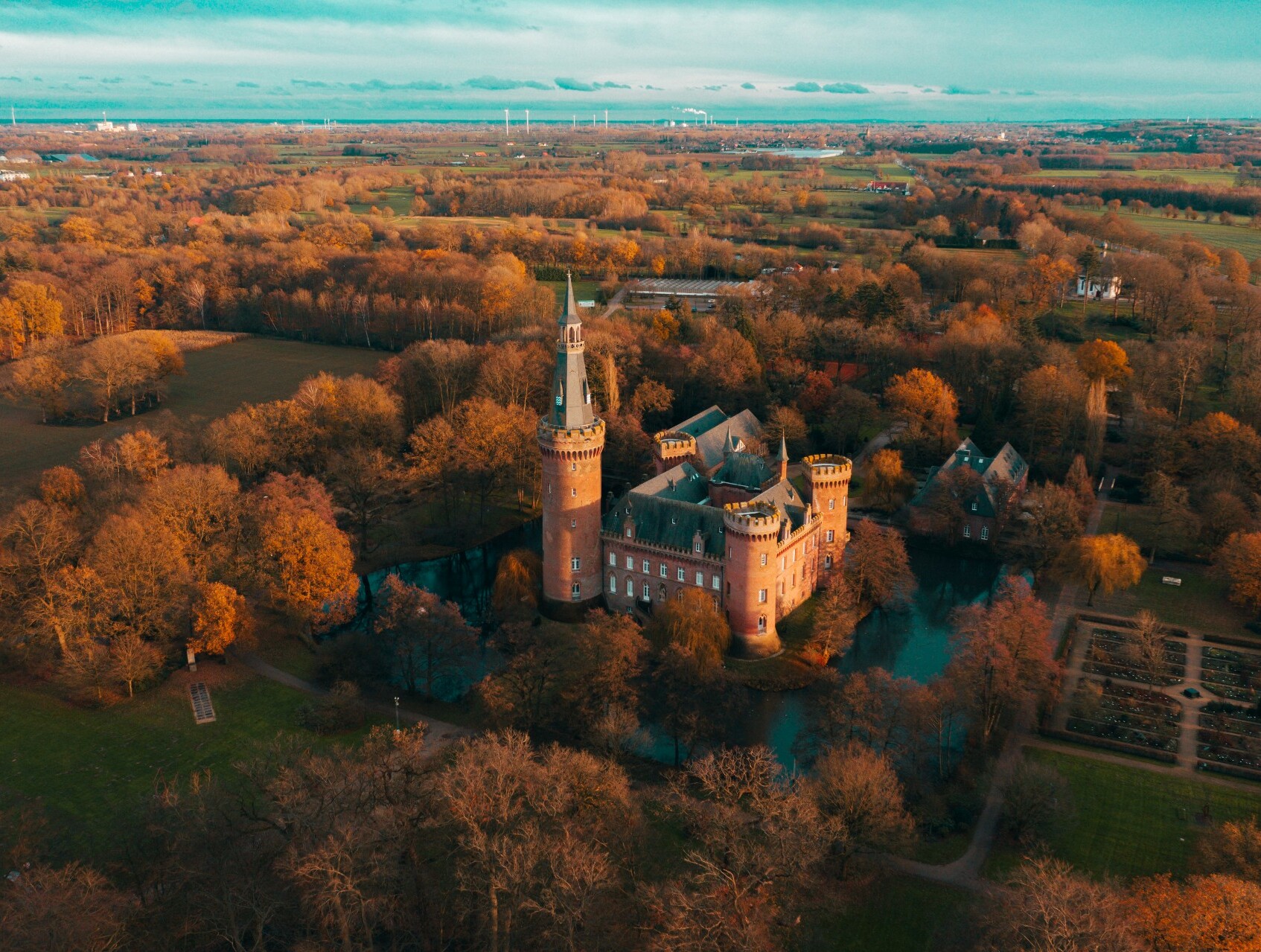 Drohnenaufnahme von Schloss Moyland in Bedburg-Hau am Niederrhein, umgeben von in rotes Licht getauchten Wäldern und Feldern