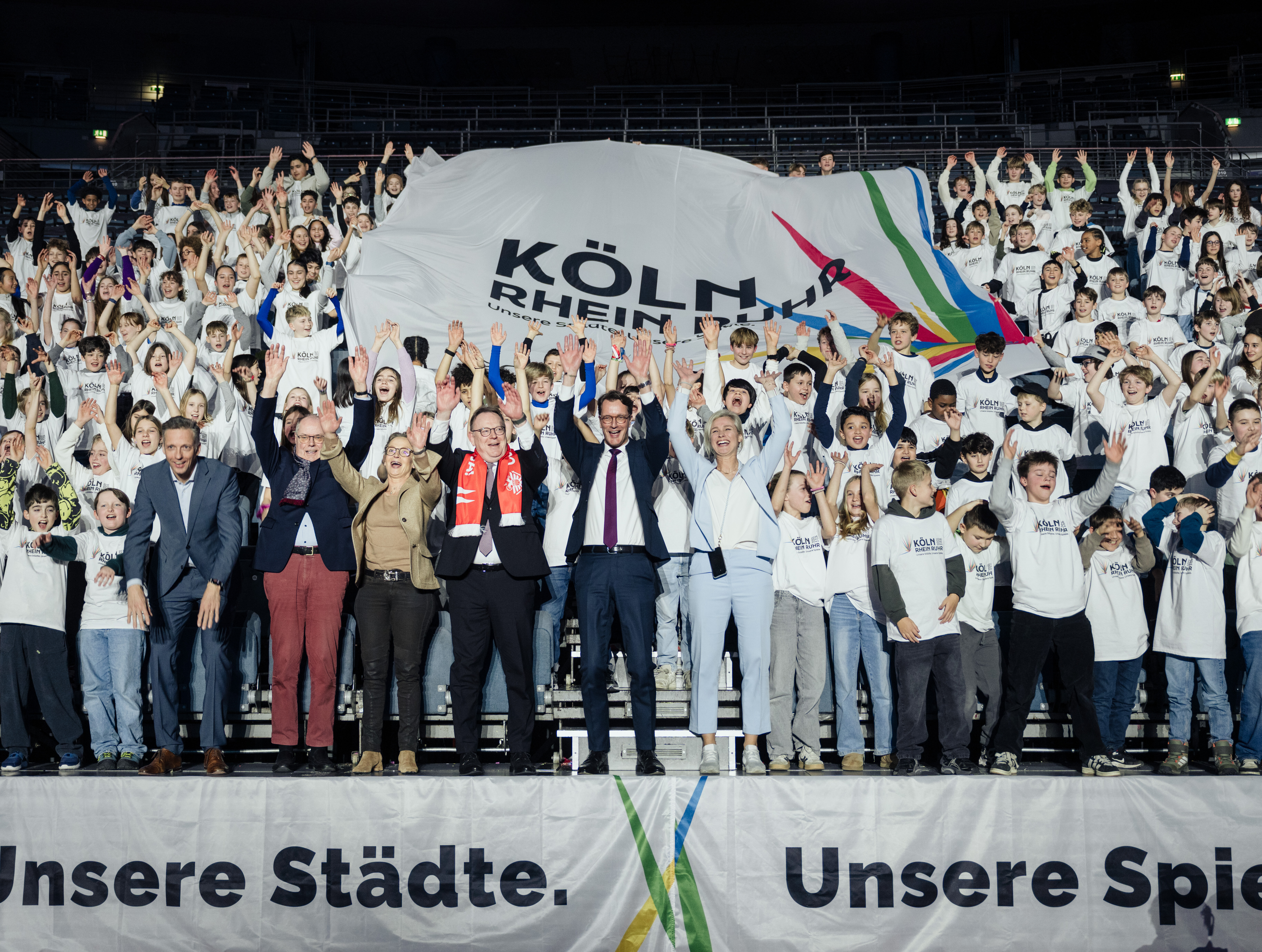 Menschen reißen bei der Auftaktveranstaltung der Bewerbungskampagne um die Olympischen und Paralympischen Spiele an Rhein und Ruhr in der Lanxess-Arena die Arme in die Luft und halten einen Banner mit dem Schriftzug KölnRheinRuhr 