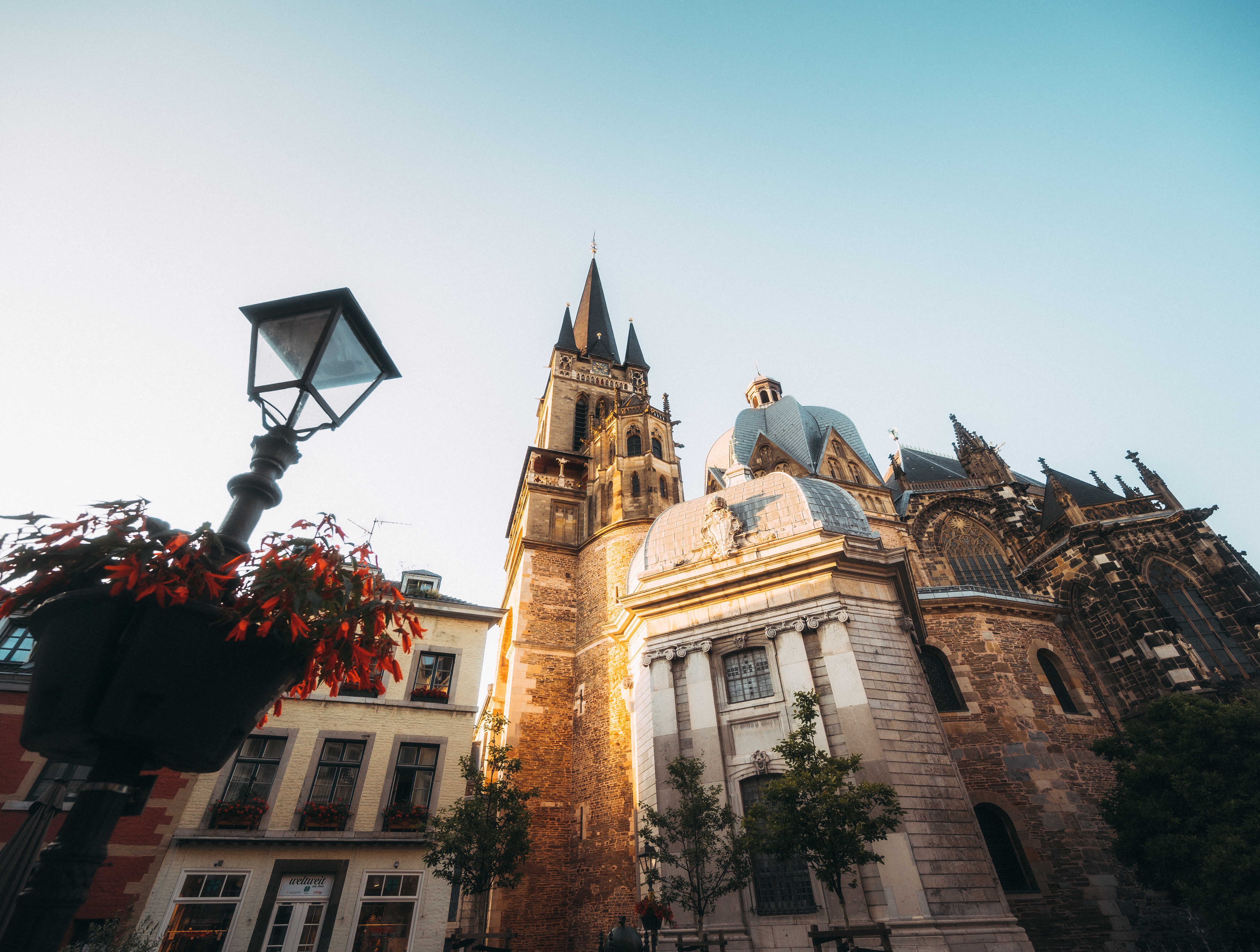 Seitenansicht des Aachener Doms mit historischer Laterne und Blumenampel, von unten fotografiert. 