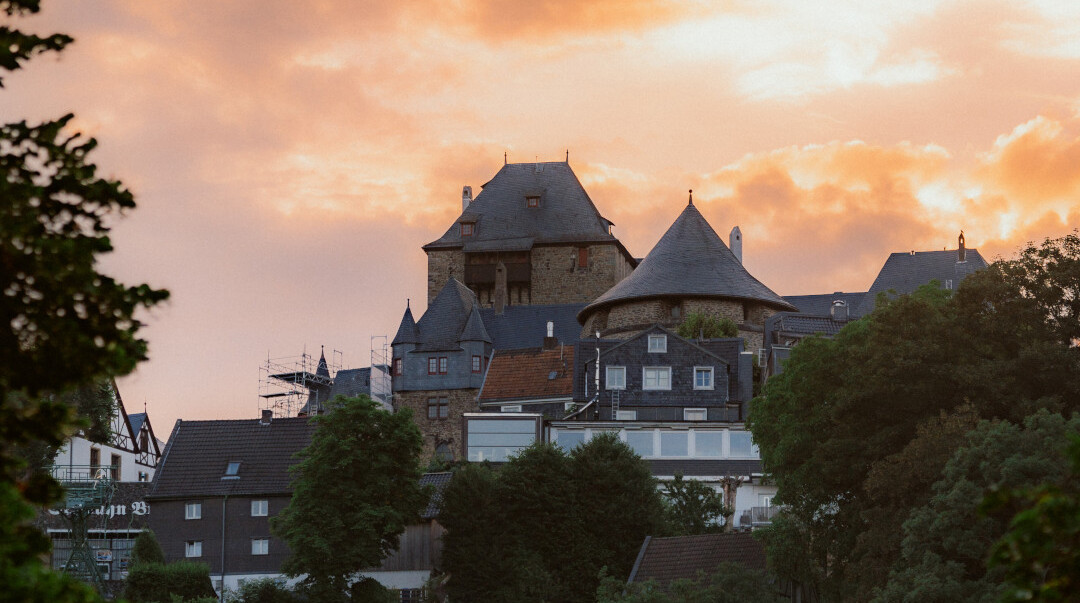 Schloss Burg in Solingen, vor einem Himmel mit orange leuchtenden Wolken bei Sonnenuntergang, umgeben von Häusern und Bäumen.