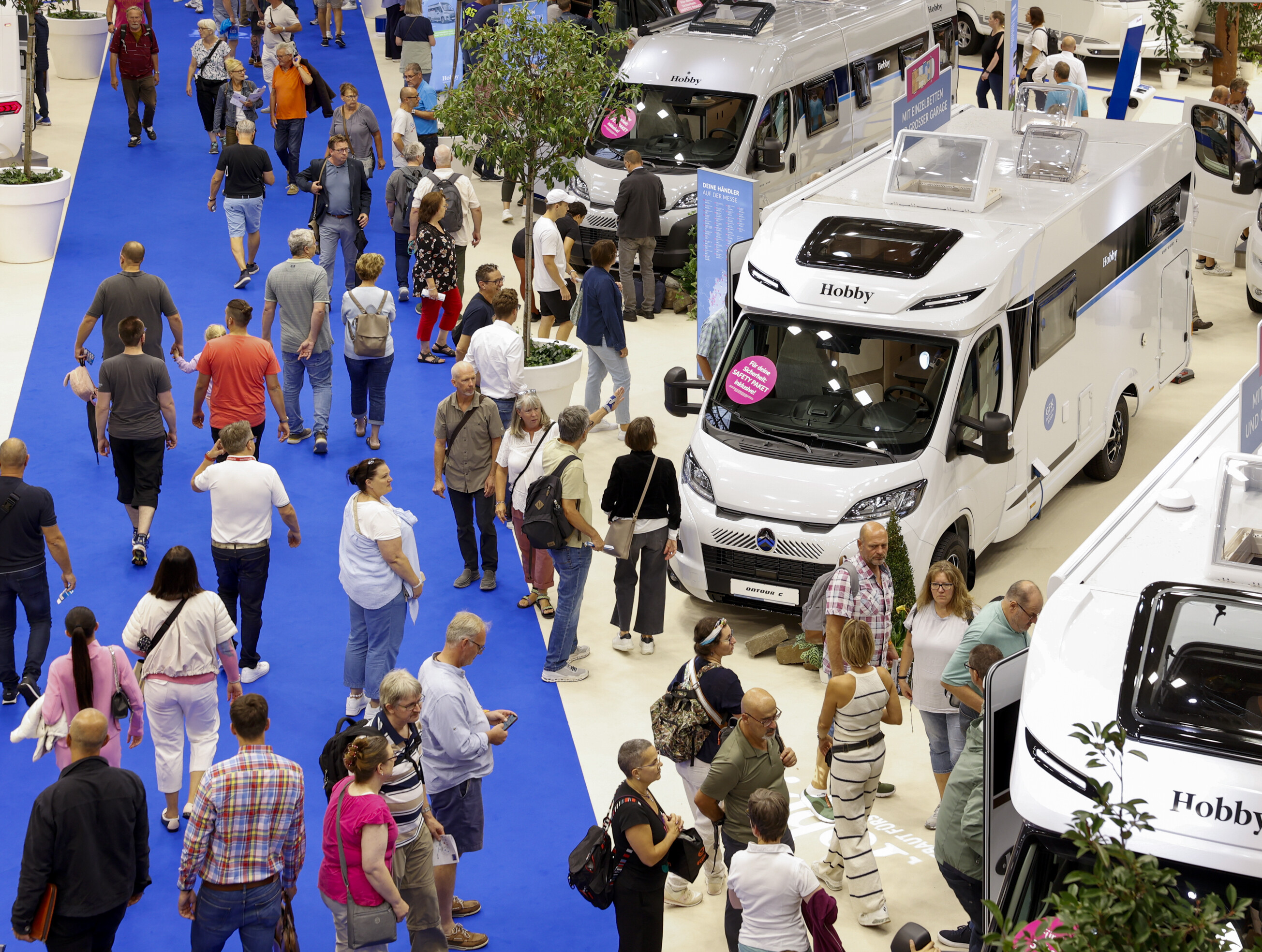 Menschenmenge auf einem blauen Teppich in einer Messehalle der Messe Düsseldorf beim Caravan Salon 2024. umgeben von Wohnmobilen. Besucher betrachten die Fahrzeuge und stehen teilweise in Schlangen, um sie von innen zu besichtigen. 