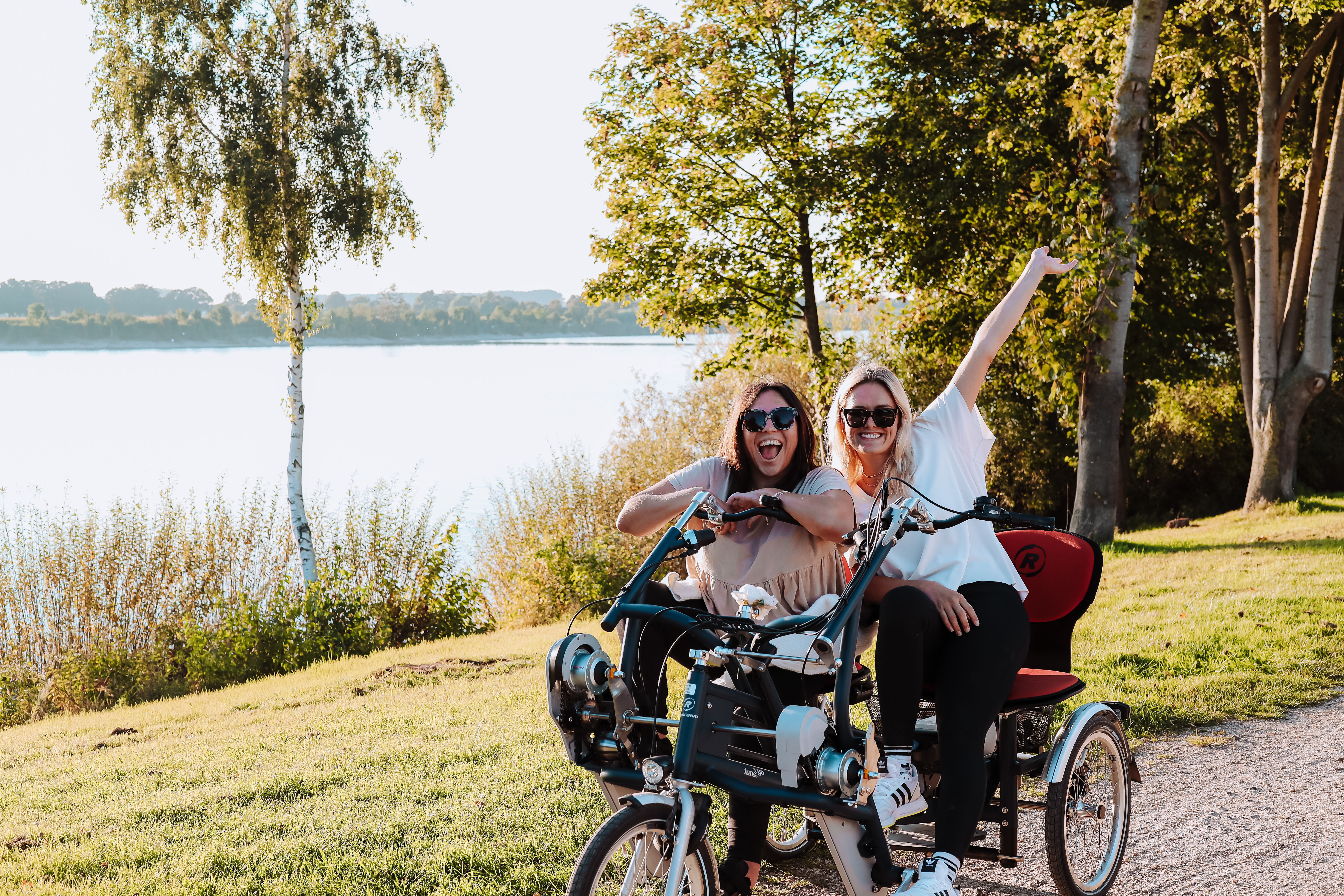 Zwei Fauen auf einem Doppel-Sitzer-Fahrrad für Rollstuhlfahrende auf einem Weg am Seeufer in Xanten. 