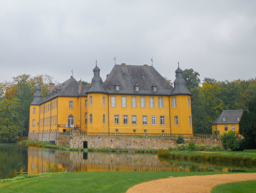 Blick auf das historische Schloss Dyck in Jüchen am Niederrhein, umgeben von einem weitläufigen Schlosspark. Das gelb gestrichene Wasserschloss mit seinen markanten Türmen spiegelt sich im ruhigen Wasser des Schlossgrabens wider. Im Vordergrund sind gepflegte Rasenflächen und Wege zu sehen, die von üppiger Vegetation und hohen Bäumen umgeben sind.