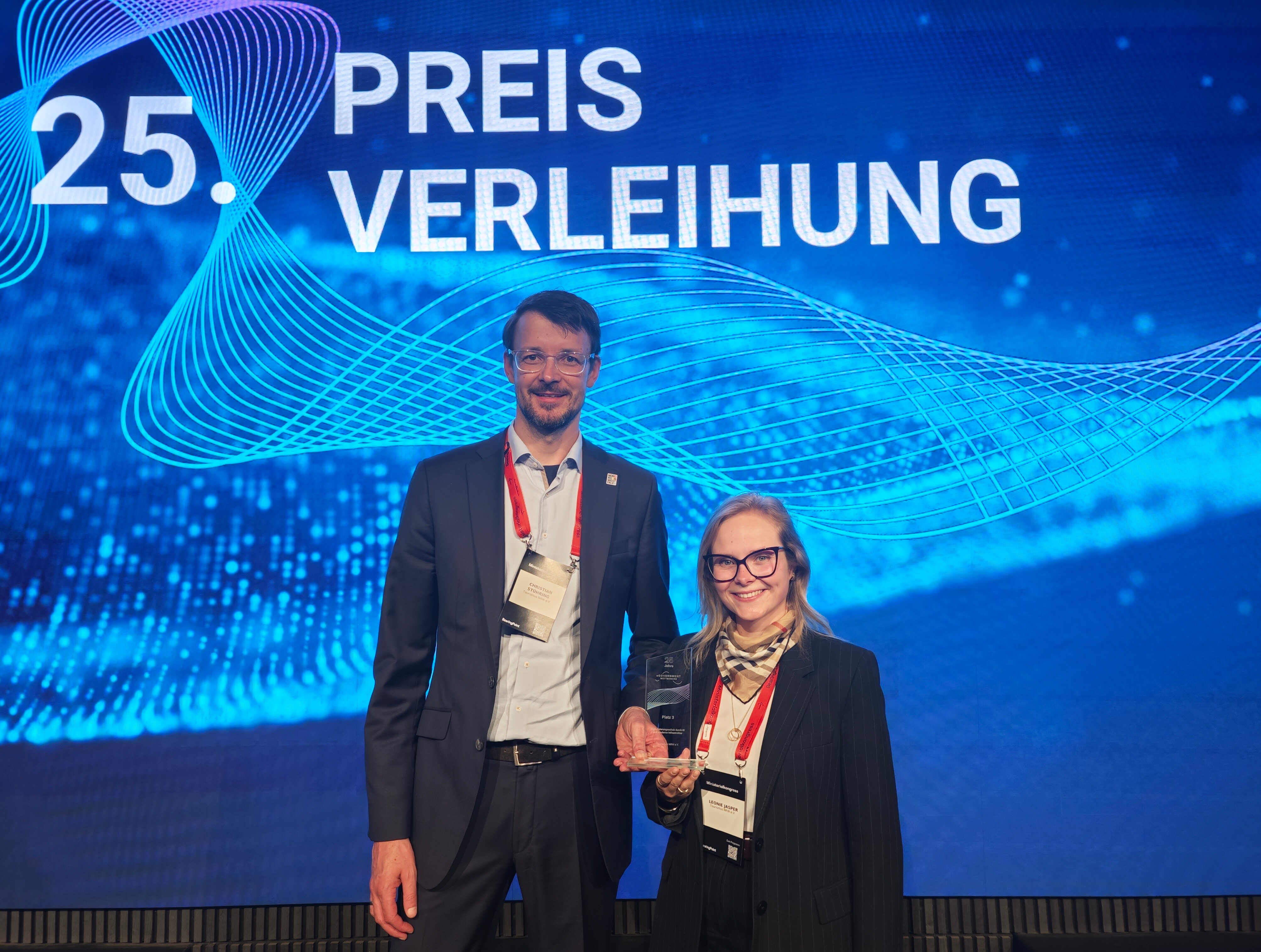 Zwei Mitarbeitende von Tourismus NRW halten den Glas-Award bei der Preisverleihung des eGovernment-Wettbewerbs 2026.  
