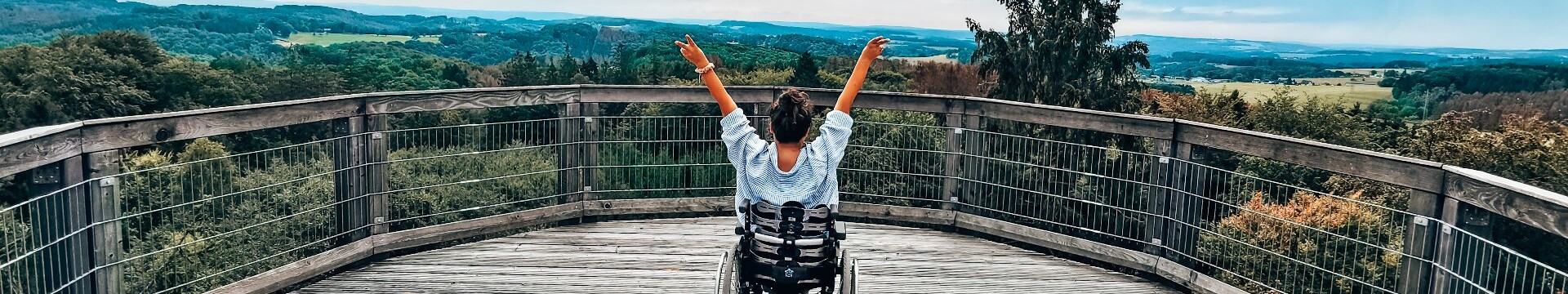 Eine Rollstuhlfahrerin schaut von der Aussichtsplattform des Baumwipfelpfads Panabora auf die grünen Hügel im Bergischen Land und recht die Arme in die Höhe.  © wheeliewanderlust
