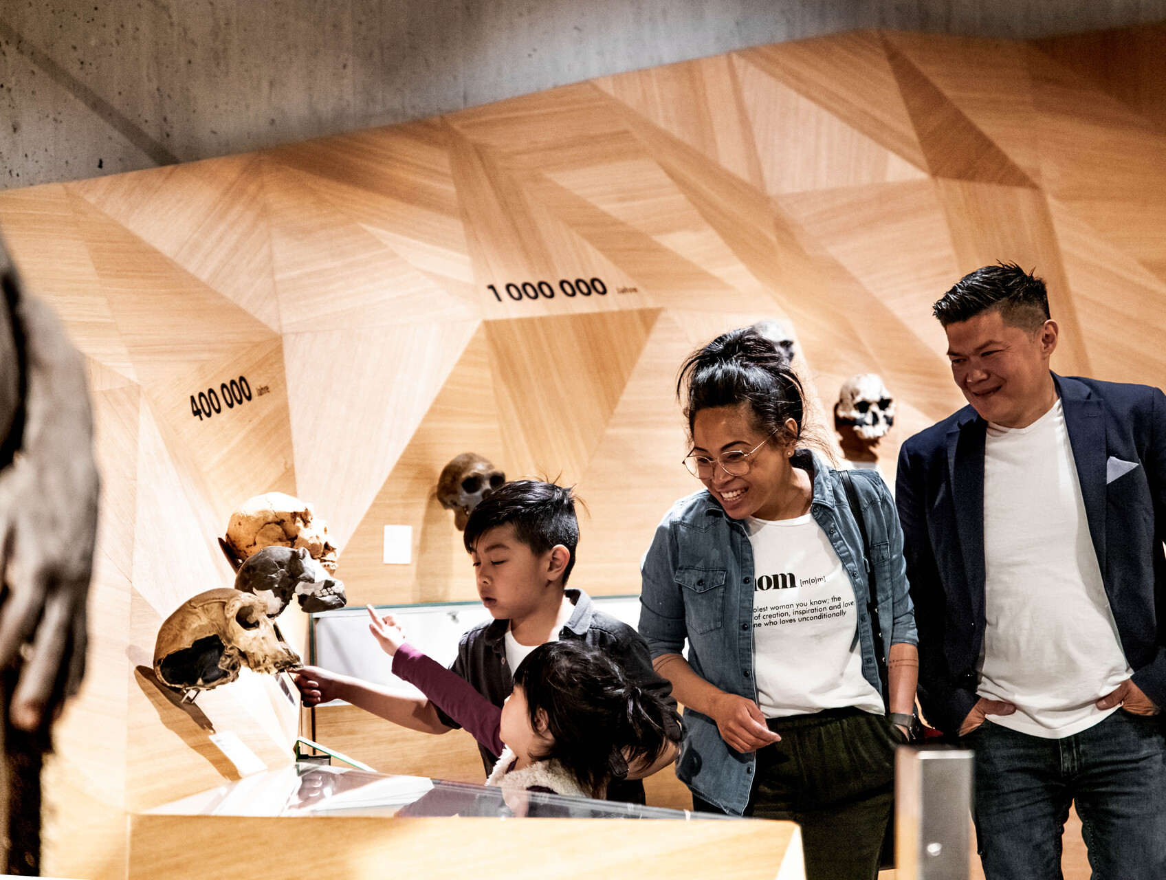 Foto einer Familie im Neanderthal Museum. Im Vordergrund betrachten zwei Kinder interessiert mehrere Schädelmodelle, die an einer Wand mit Holzmuster angebracht sind. Die Wand zeigt Zeitleisten mit den Zahlen „400.000 Jahre“ und „1.000.000 Jahre“. Das ältere Kind zeigt auf einen Schädel, während das jüngere ebenfalls darauf deutet. Dahinter stehen eine Frau mit Brille, die lächelnd zusieht, und ein Mann im dunkelblauen Sakko, der ebenfalls lächelt. Am linken Bildrand ist ein Teil einer menschlichen Figur zu sehen.