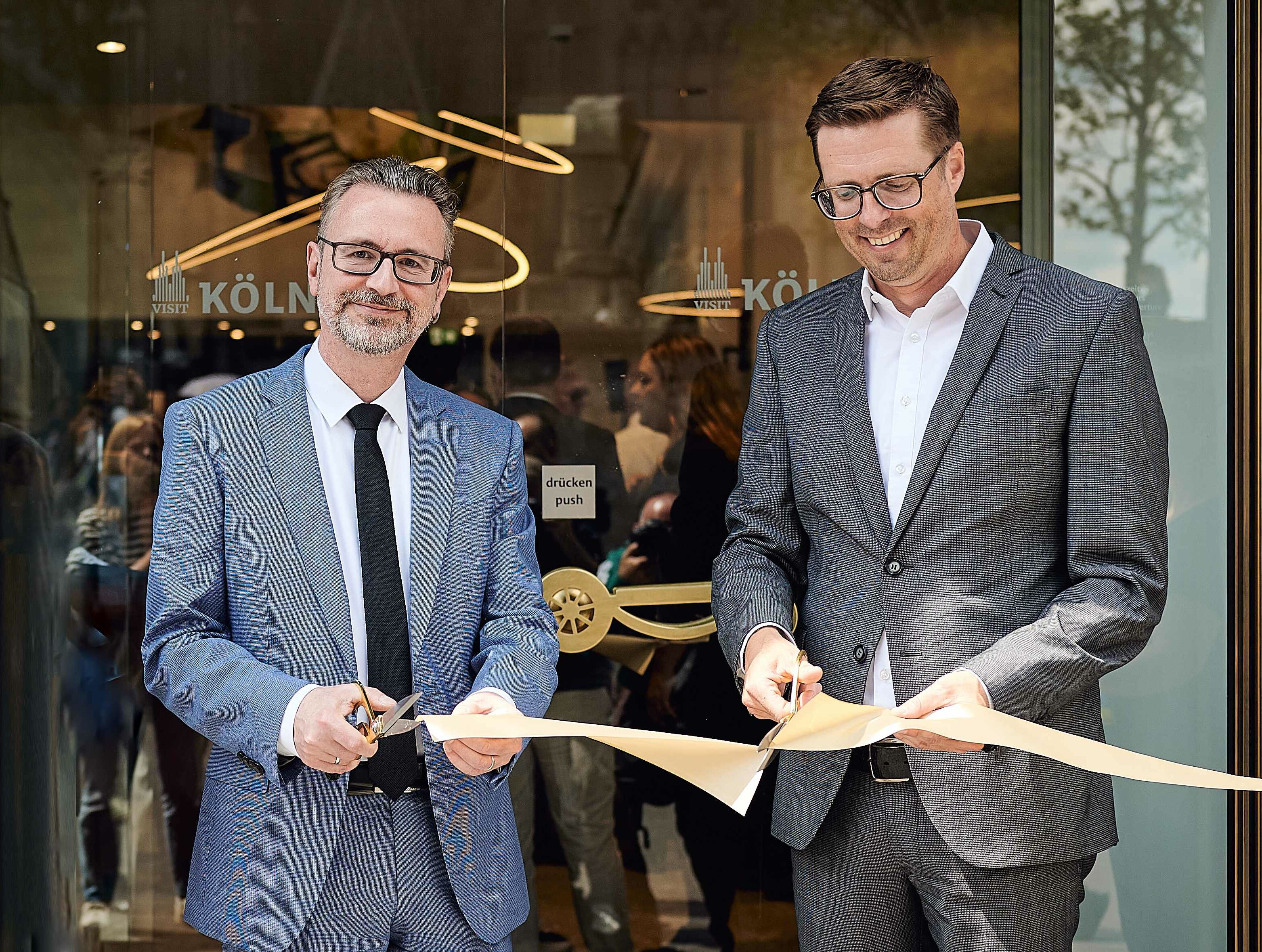 Der Geschäftsführer von KölnTourismus, Dr. Jürgen Amann, und Kölns Wirtschaftsdezernent Andree Haack, schneiden ein Band vor der Tourist-Info am Kölner Dom durch. 