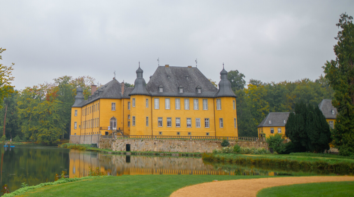 Blick auf das historische Schloss Dyck in Jüchen am Niederrhein, umgeben von einem weitläufigen Schlosspark. Das gelb gestrichene Wasserschloss mit seinen markanten Türmen spiegelt sich im ruhigen Wasser des Schlossgrabens wider. Im Vordergrund sind gepflegte Rasenflächen und Wege zu sehen, die von üppiger Vegetation und hohen Bäumen umgeben sind.