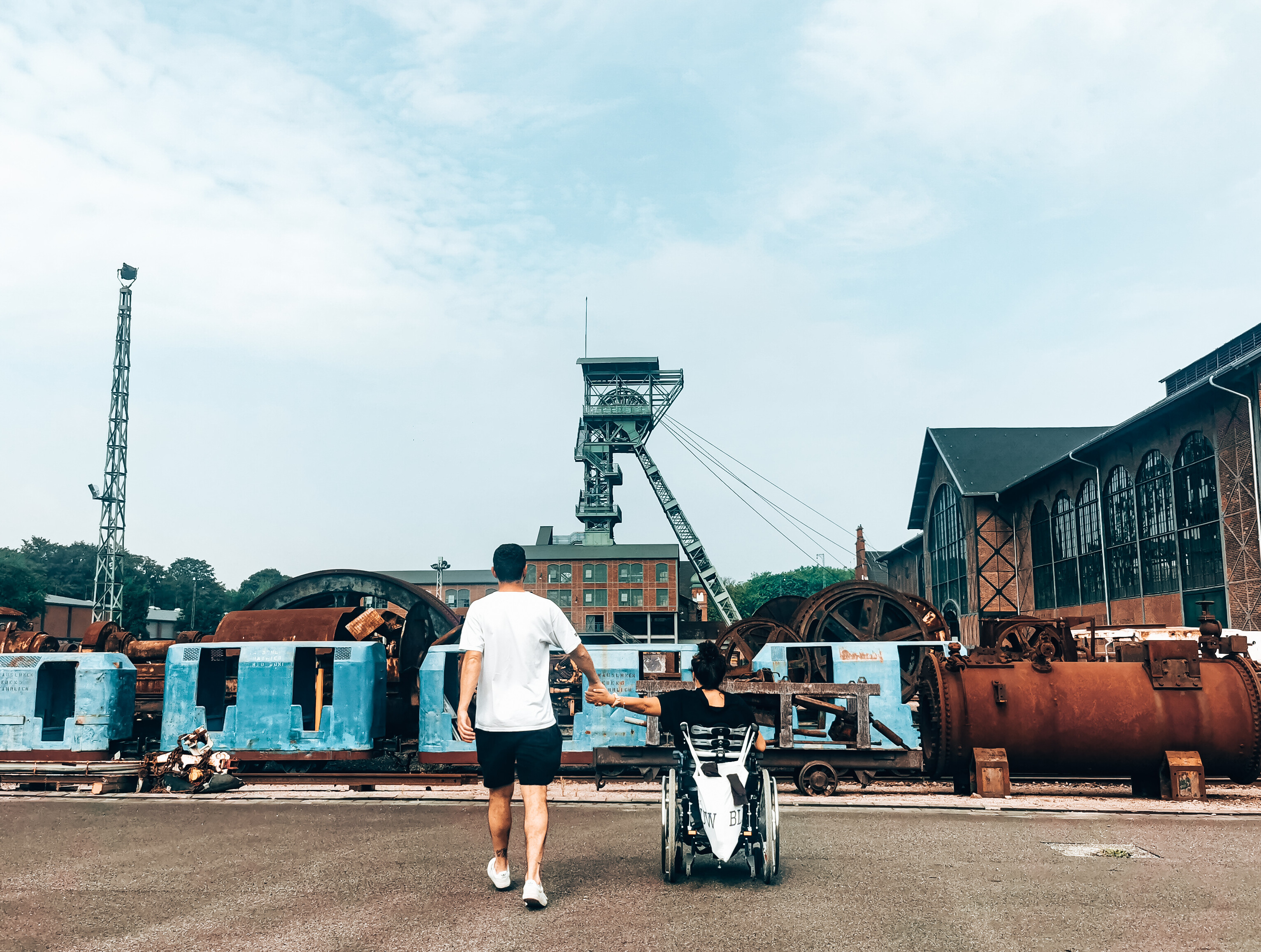 Eine Frau im Rollstuhl und ein Mann schauen auf das Gelände des Industriedenkmals Zeche Zollern in Dortmund. Im Hintergrund sind der ehemalige Förderturm und Backsteinfassade der Maschinenhalle zu sehen. © wheeliewanderlust