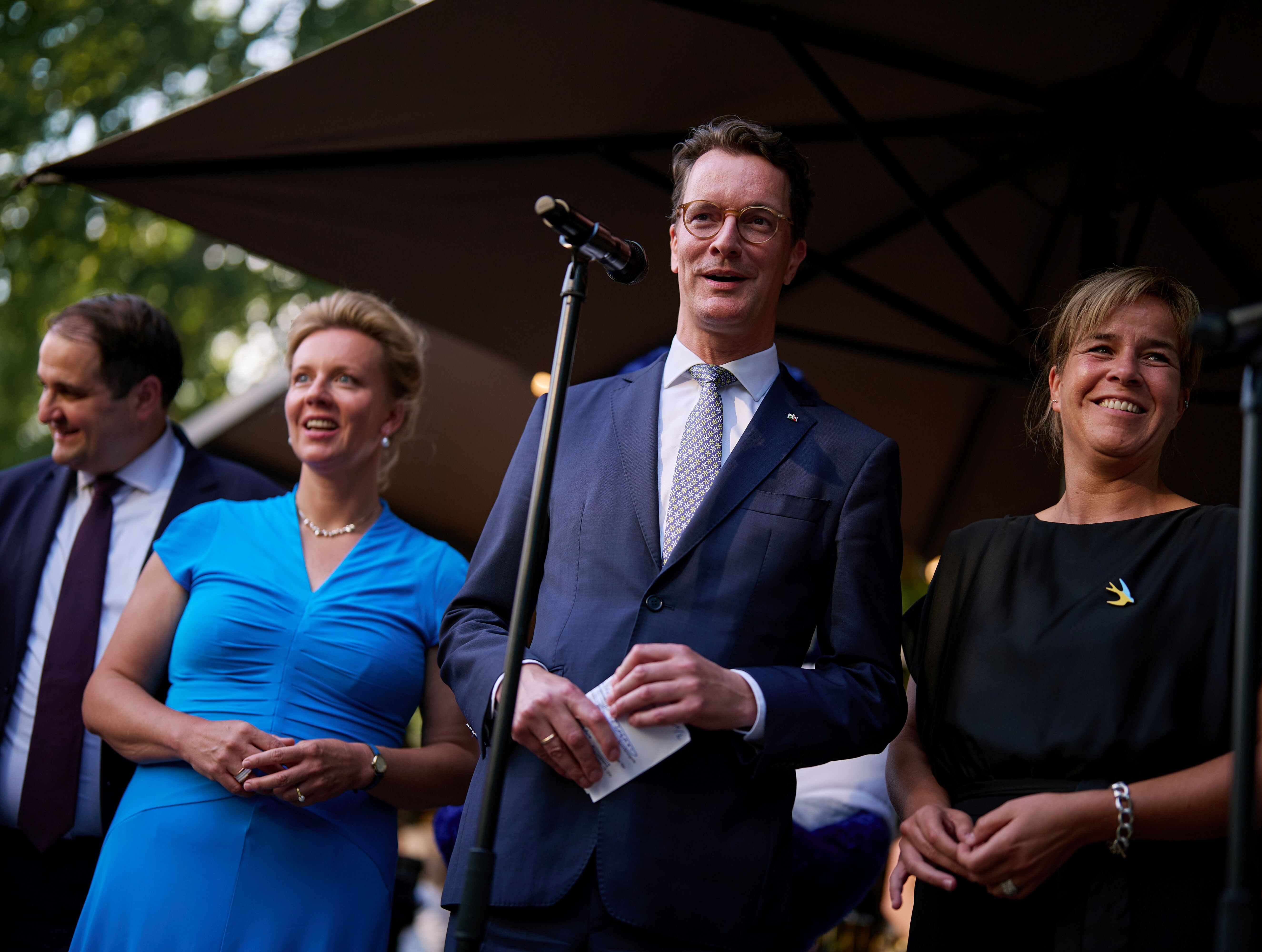 NRW-Ministerpräsident Hendrik Wüst auf dem Sommerfest der NRW-Landesregierung in Berlin 2025 © NRW-Landesvertretung NRW-Ministerpräsident Hendrik Wüst steht beim Sommerfest der NRW-Landesregierung in Berlin vor einem Mikrofon. Neben ihm stehen NRW-Wirtschaftsministerin Mona Neubaur und NRW-Kulturministerin Ina Brandes.