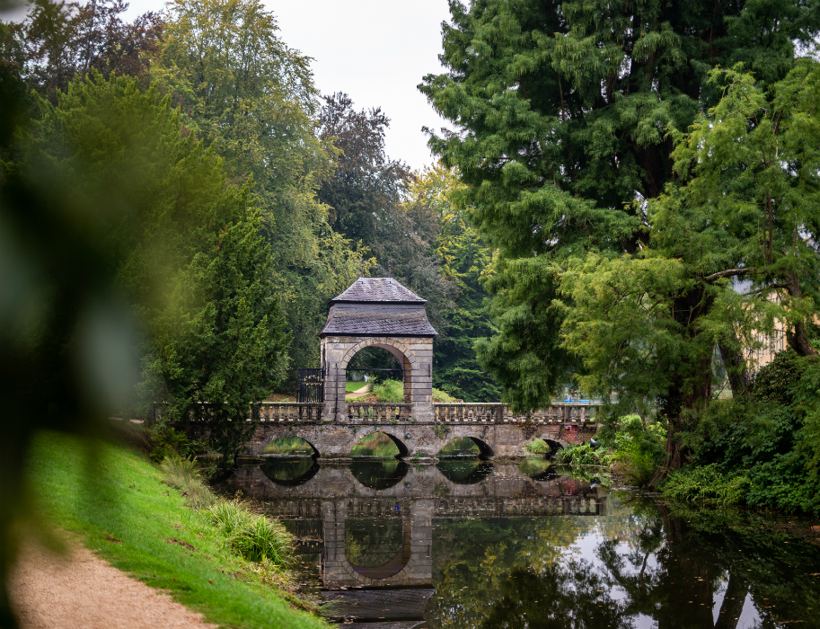 Ein steinernes Torhaus steht im Park von Schloss Dyck in Jüchen am Niederrhein auf einer Brücke, die ein ruhiges Gewässer überquert. Die Brücke hat mehrere Bögen, die sich im Wasser spiegeln. Das Torhaus ist von dichtem Grün umgeben, darunter große Bäume und Büsche. 