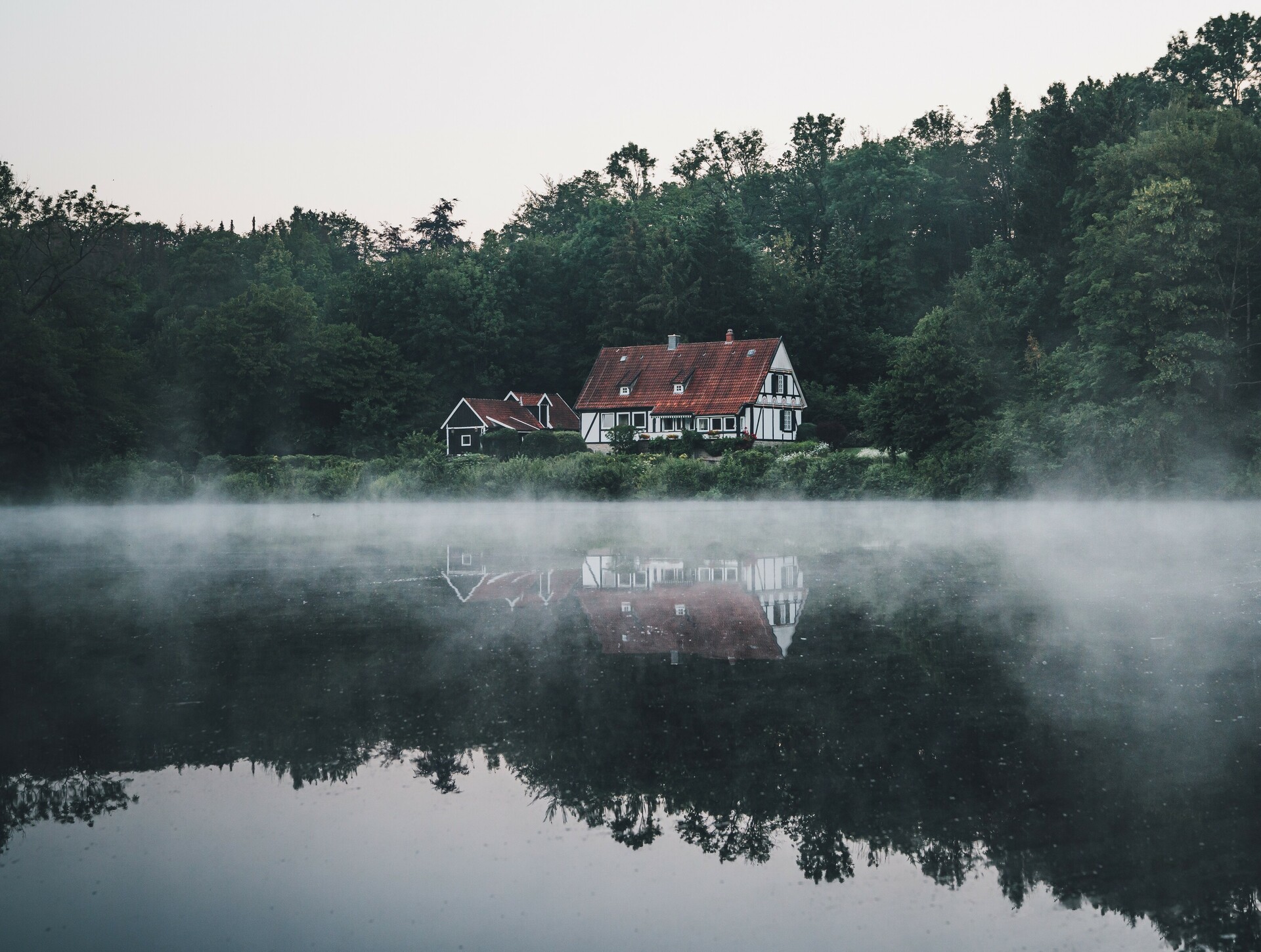 Am unteren Teich an den Externsteinen spiegelt sich ein Fachwerkhaus im Wasser, über dem noch leichte Nebelschwaden liegen. 