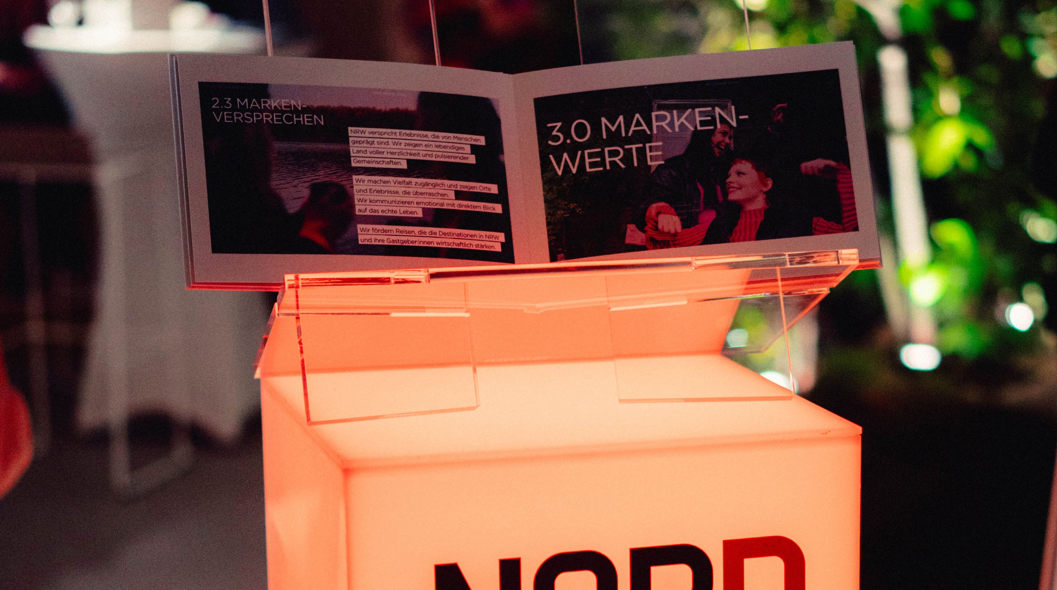 Neuer Markenauftritt von DeinNRW © Thomas Kaup/RP Creatives, Tourismus NRW e.V. Auf einem erleuchteten Markenlogo von DeinNRW steht ein aufgeklapptes Brand Book zum neuen Markenauftritt von DeinNRW.