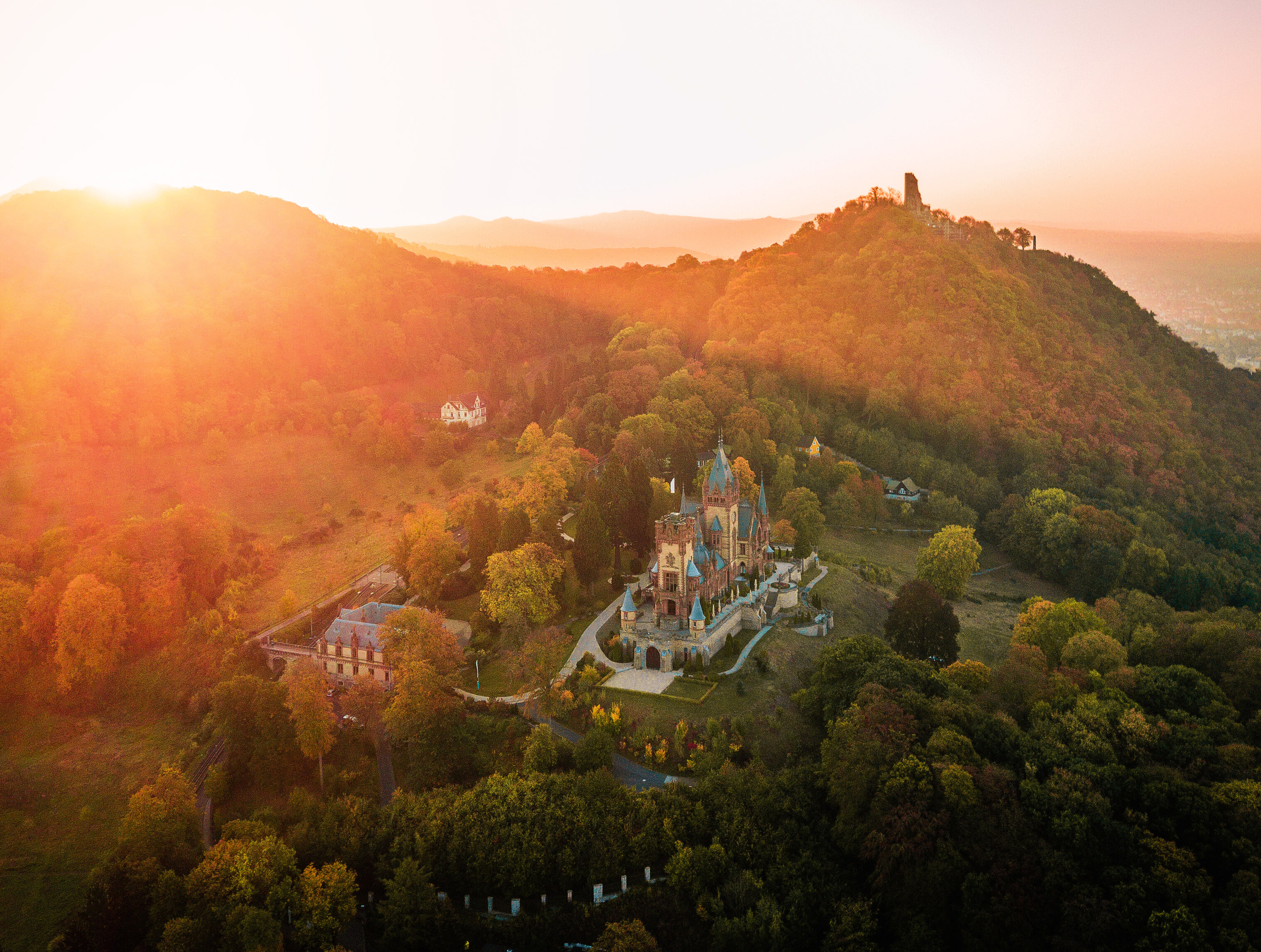 Blick auf das in rötliches Morgenlicht getauchte Schloss Drachenburg bei Königswinter, umgeben von bewaldeten Hügeln im Naturpark Siebengebirge 