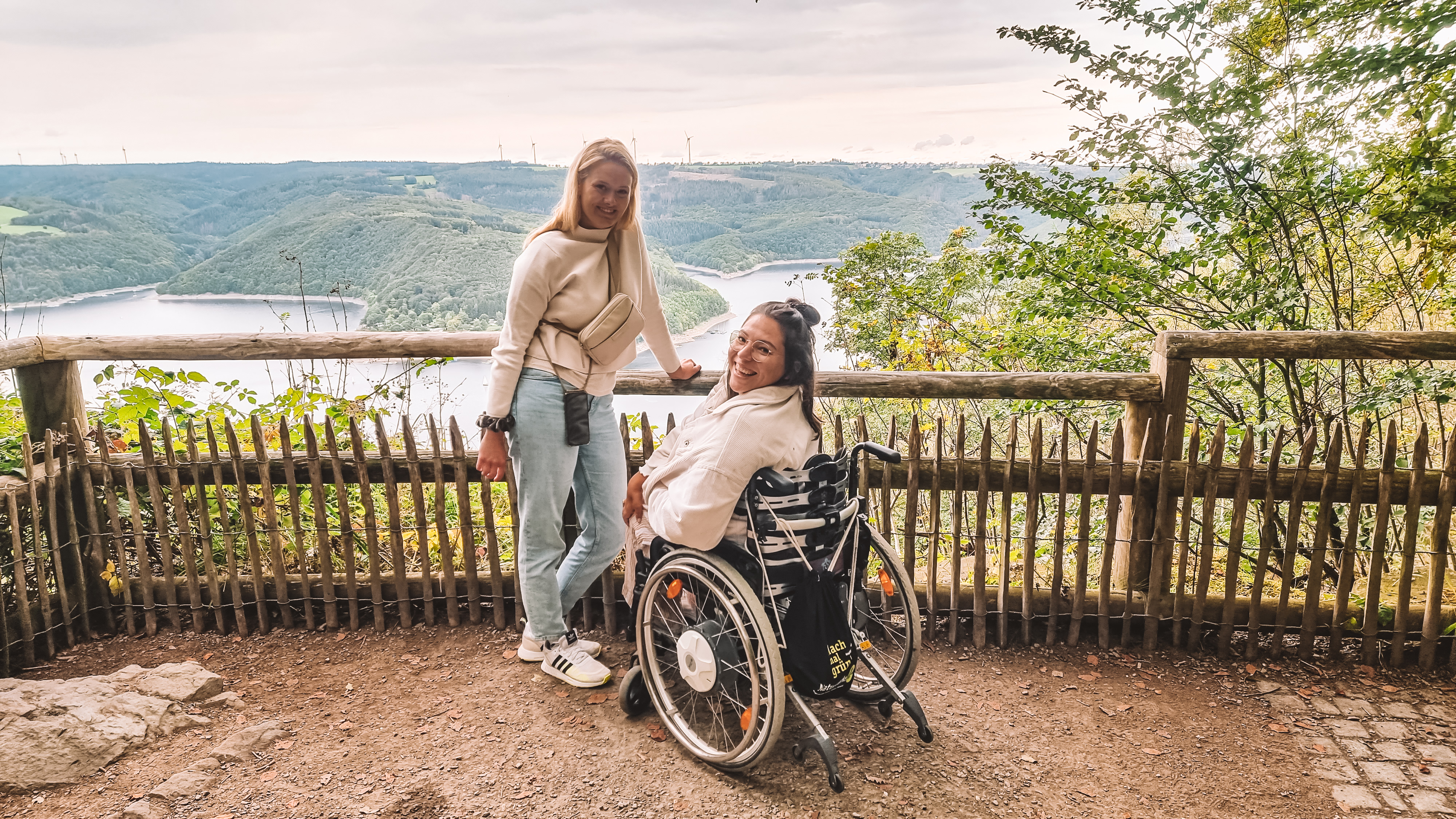 Eine Frau im Rollstuhl und eine zweite Frau vor dem Zaun einer Aussichtplattform mit Blick auf die Wälder und Seen im Nationalpark Eifel. 