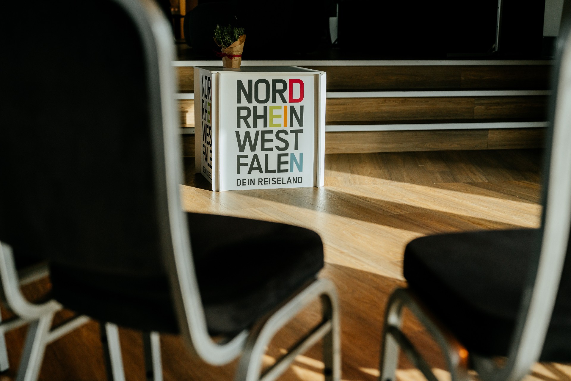 Foto eines weißen, würfelförmigen Aufstellers mit der Aufschrift „Nordrhein-Westfalen – Dein Reiseland“ in farbigen Buchstaben, auf einem hölzernen Boden in einem Veranstaltungsraum. Oben auf dem Würfel steht ein kleiner Topf mit einer Pflanze. Im Vordergrund sind unscharf die Lehnen und Sitzflächen von schwarzen Stühlen zu sehen. Im Hintergrund befindet sich eine hölzerne Bühne mit Stufen, die von Licht und Schatten durchzogen ist.
