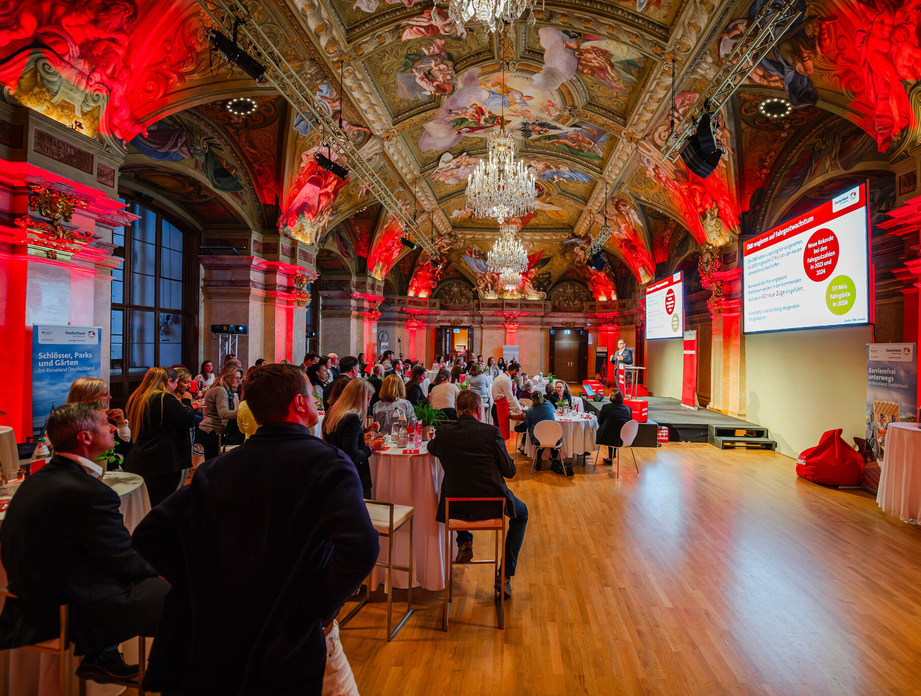 DZT-Roadshow durch Wien und Linz © DZT/Kamila Stojanonová Reiseanbietende, Medienschaffende und Blogger:innen sitzen bei der DZT-Roadshow in Österreich in einem prächtigen Barocksaal mit Deckengemälden und roter Beleuchtung.