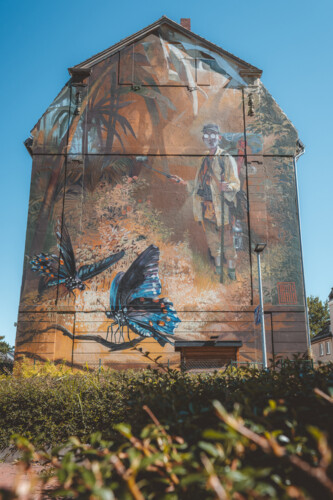 Ein Mural von Case Maclaim in Gelsenkirchen-Ückendorf zeigt große Schmetterlinge und einen Mann, der einen Selfie-Stick in der Hand hält. Es ist eine Neuinterpretation des Gemäldes "Der Schmetterlingsfänger" von Carl Spitzweg.