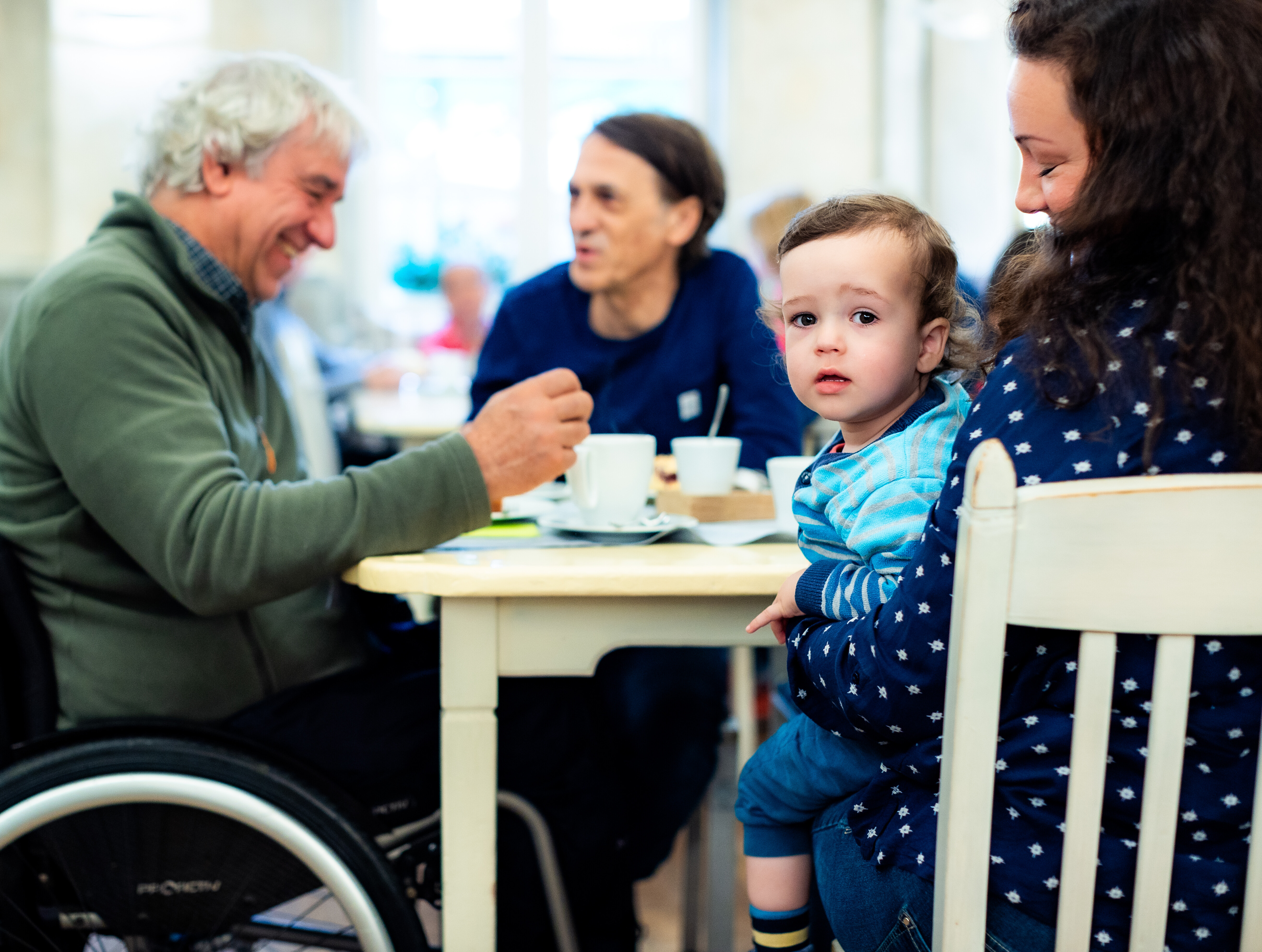 Älterer Mann mit grauen Haaren im Rollstuhl lacht und unterhält sich mit einem Mann mittleren Alters an einem Café-Tisch. Eine Frau mit braunen Locken hält ein Kleinkind auf dem Schoß; das Kind blickt neugierig in die Kamera. Auf dem Tisch stehen Kaffeetassen und Unterteller. 