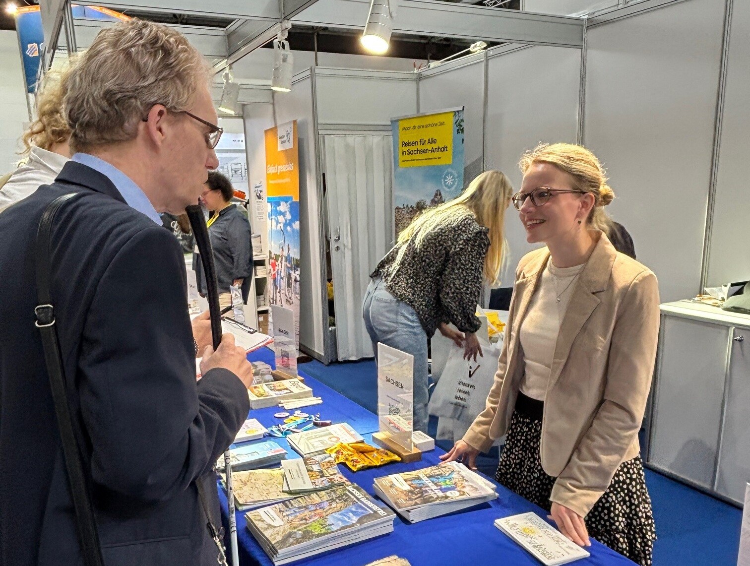 NRW-Präsenz auf der Rehacare 2026 © Tourismus NRW e.V. Eine Mitarbeiterin von Tourismus NRW ist auf der Rehacare in ein Gespräch mit einem Besucher vertieft. Vor ihr liegen Broschüren zum barrierefreien Reisen in NRW auf einem Tisch.