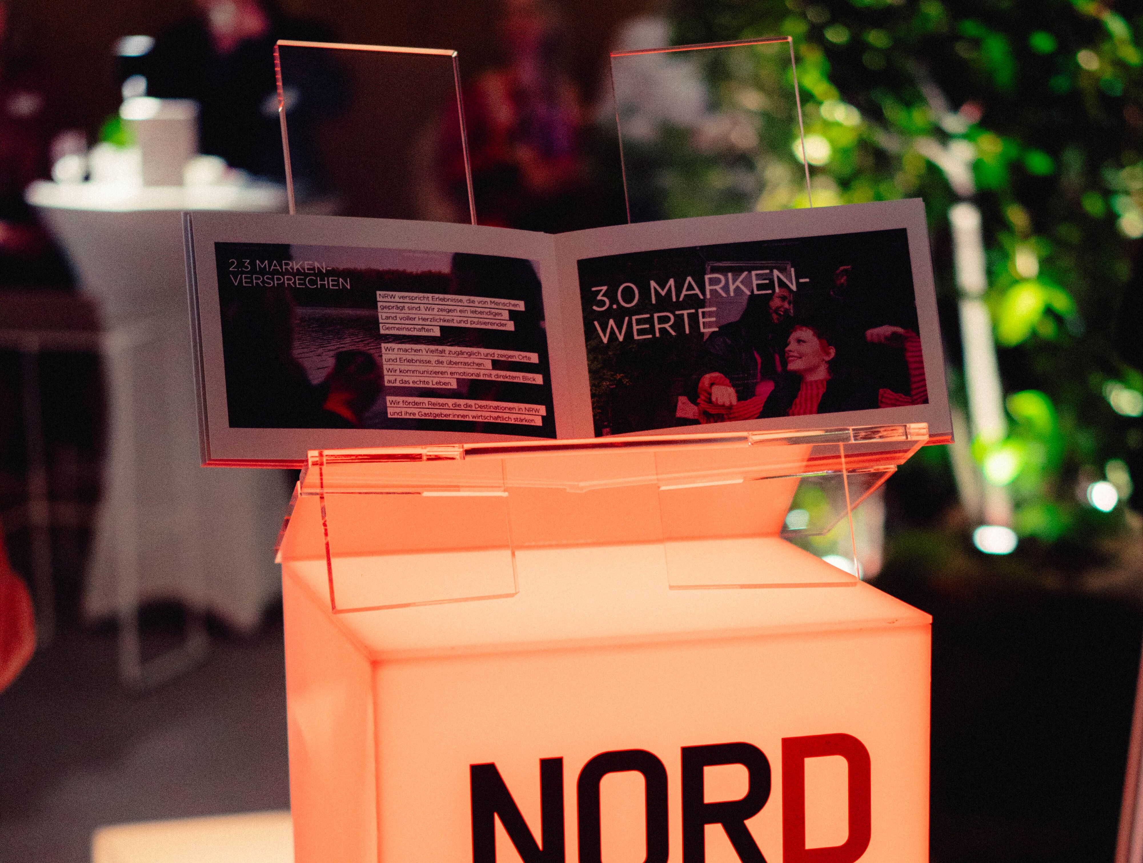 Auf einem erleuchteten Markenlogo von DeinNRW steht ein aufgeklapptes Brand Book zum neuen Markenauftritt von DeinNRW.