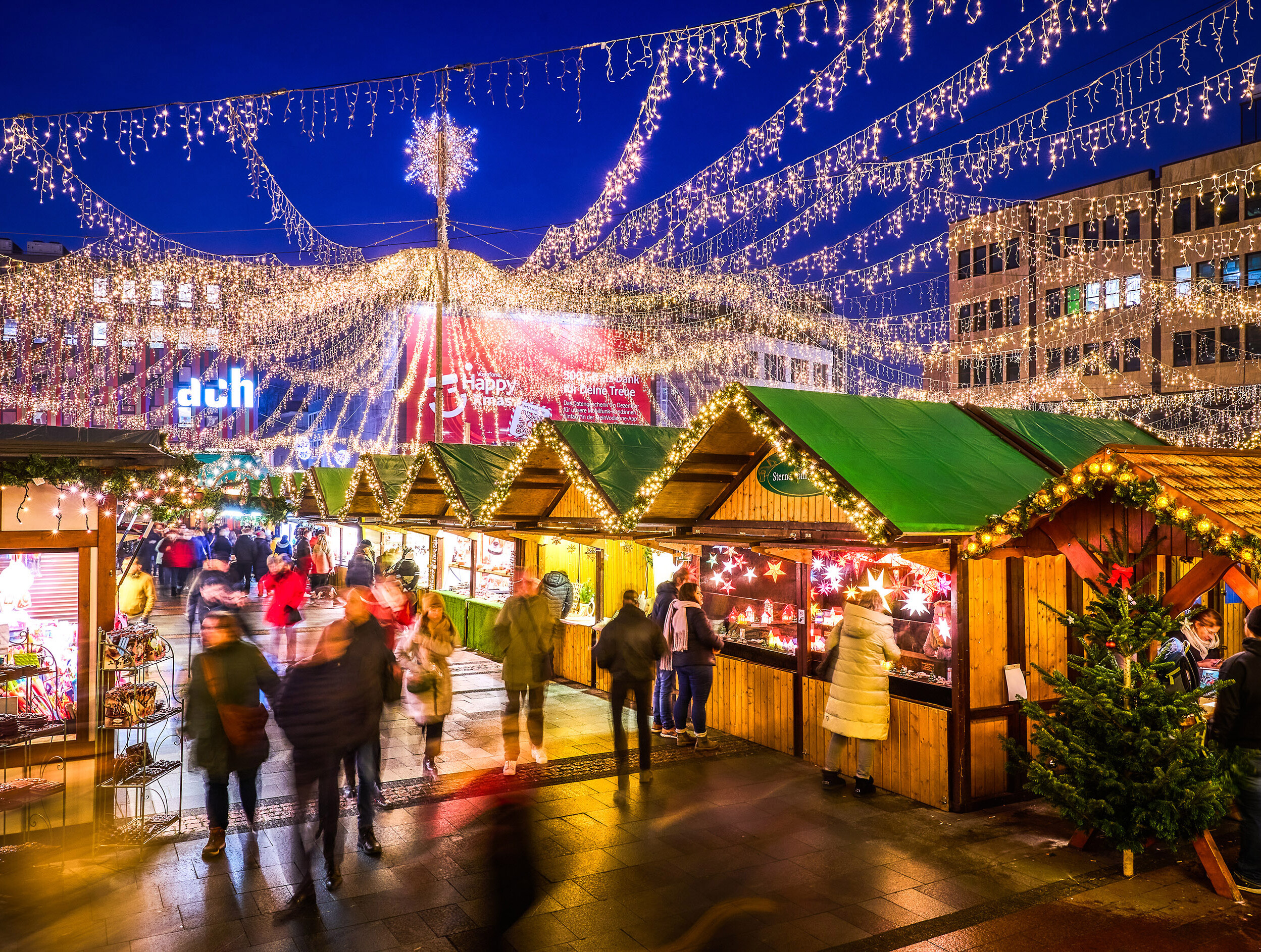 Festlich beleuchteter Internationaler Weihnachtsmarkt in Essen am Abend. Über den Holzhütten sind Lichterketten gespannt, die sich strahlenförmig von einem zentralen Punkt mit einem leuchtenden Stern aus nach außen verteilen und den Markt in ein warmes, gemütliches Licht tauchen. Besucher schlendern durch die Gänge zwischen den Buden, die verschiedene Produkte anbieten. 