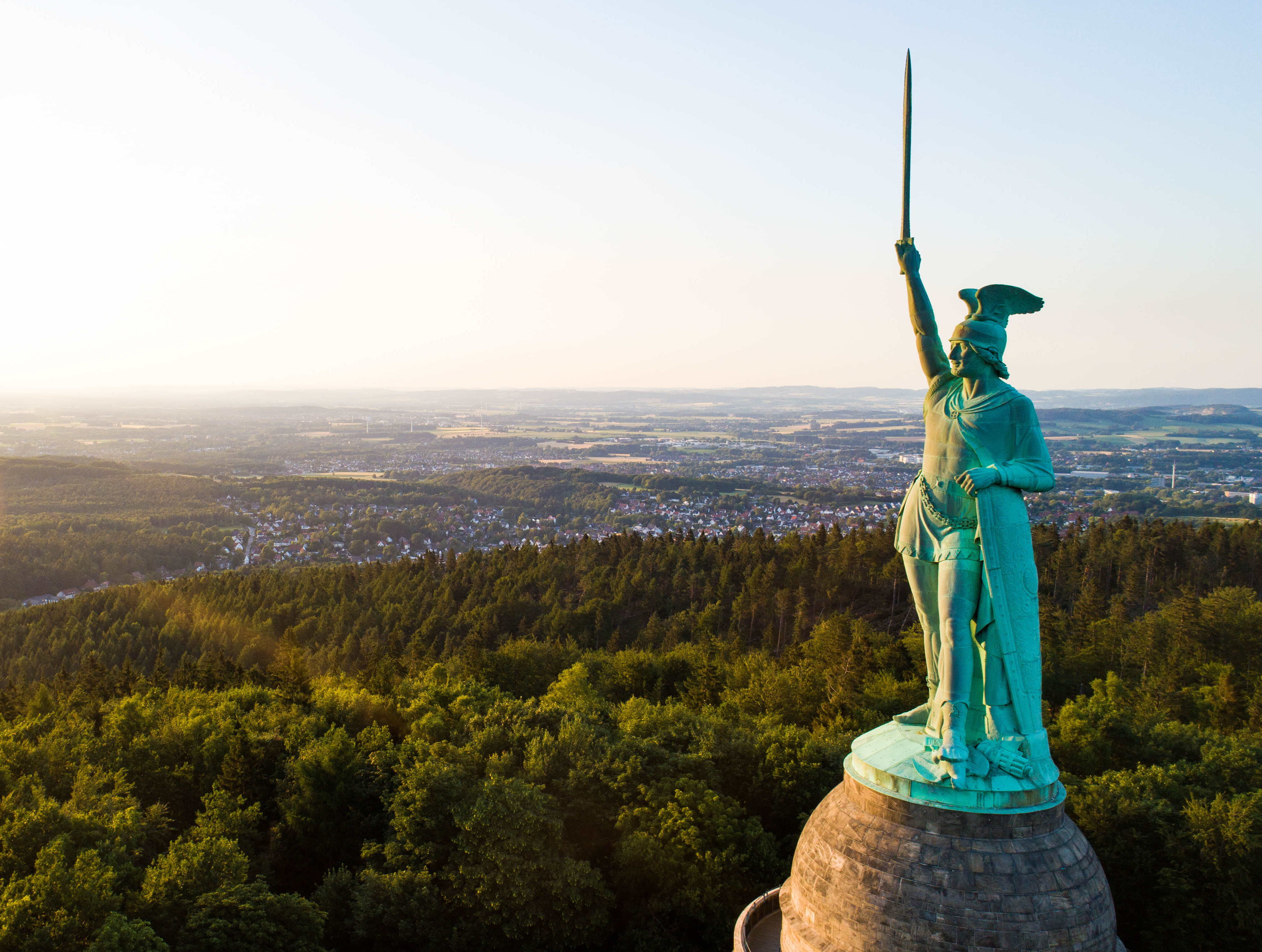 Hermannsdenkmal Teutoburger Wald © Tourismus NRW e.V. Luftaufnahme des Hermannsdenkmals im Teutoburger Wald bei Detmold in Ostwestfalen. Die grünliche Kupfer-Statue stellt Hermann, den Cherusker, dar, der einen Helm mit Flügeln trägt und ein Schwert in die Luft reckt. Die Statue steht auf einem steinernen Sockel über den Wipfeln des Teutoburger Walds.