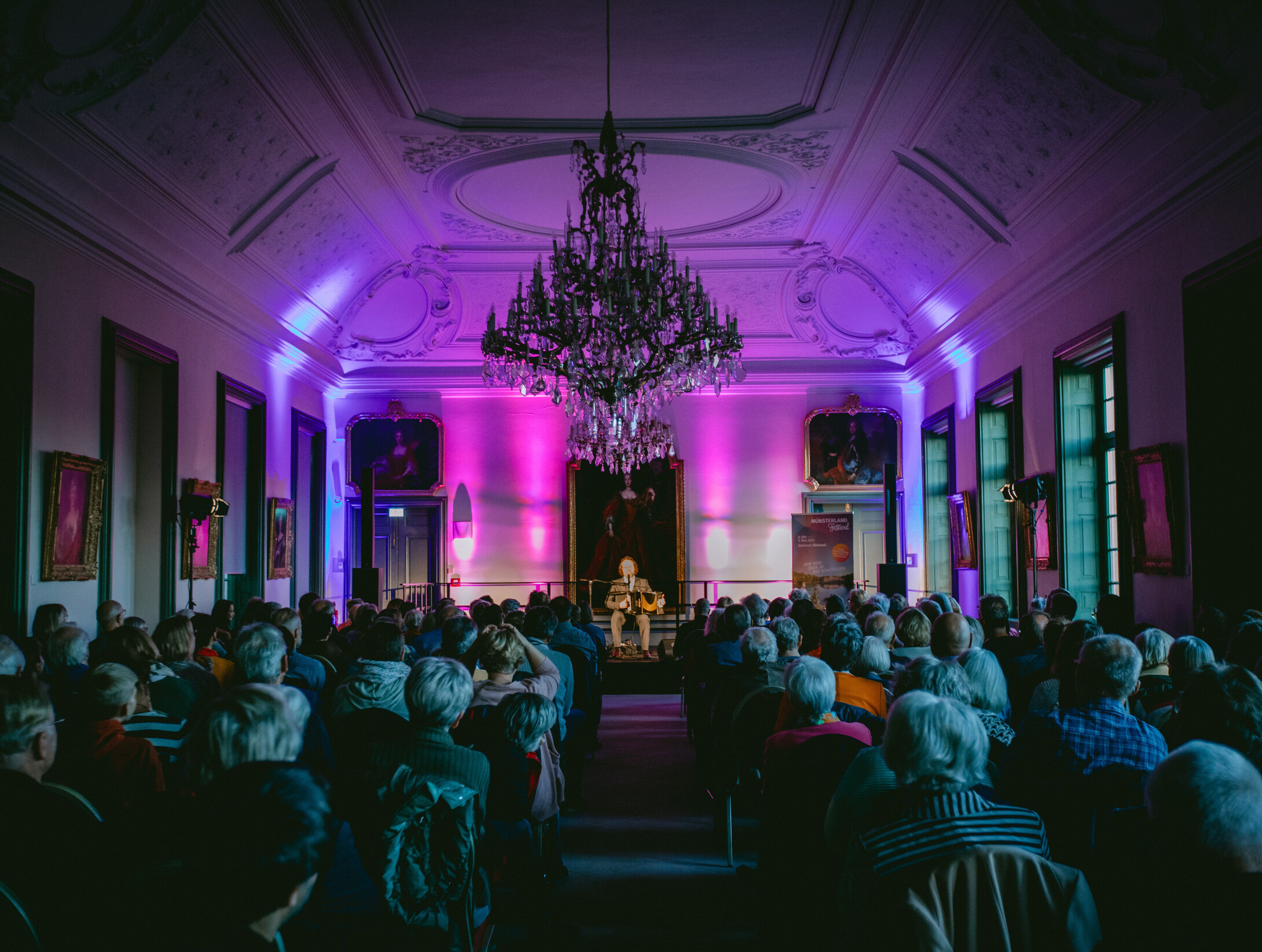 Konzert in der Oranienburg am Schloss Nordkirchen © Münsterland e.V./Maren Kuiter Ein Mann spielt auf einem kleinen Podest in einem pompösen Raum in der Oranienburg am Schloss Nordkirchen Akkordeon. An den Wänden des Raums hängen gemalte Porträts. Unter einem Kronleuchter, der an der stuckverzierten Decke hängt, sitzt das Publikum.