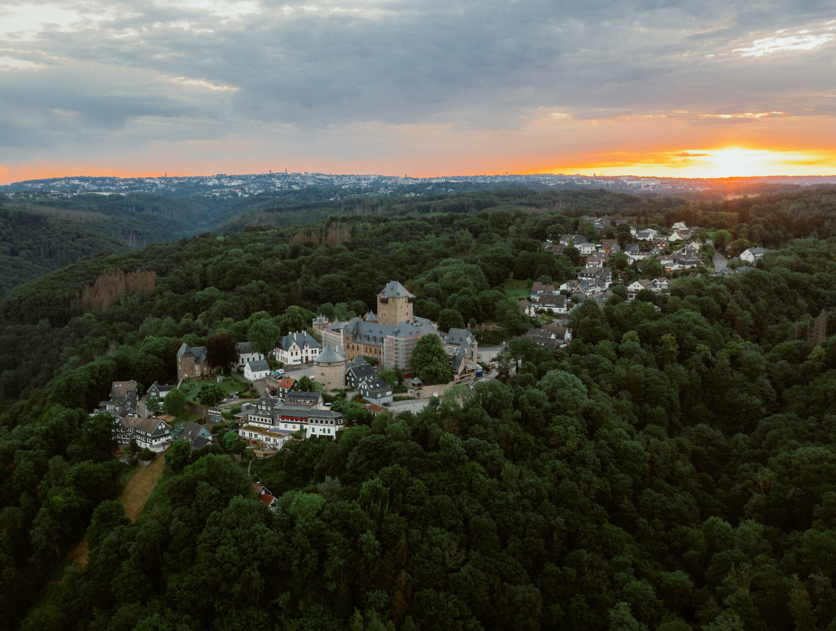 Schloss Burg umgeben von einzelnen Häusern und Wald aus der Vogelperspektive im Sonnenuntergang.