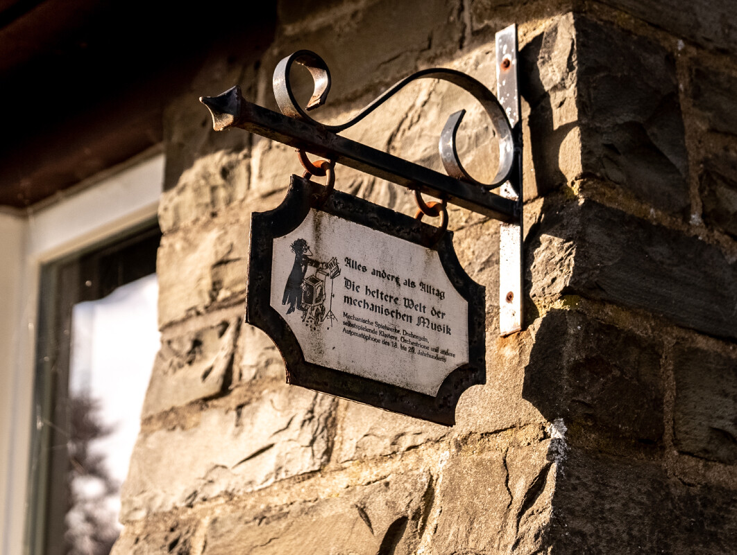 An einer Hauswand aus braunen Steinen hängt ein Schild des Bergischen Drehorgelmuseums in Marienheide.