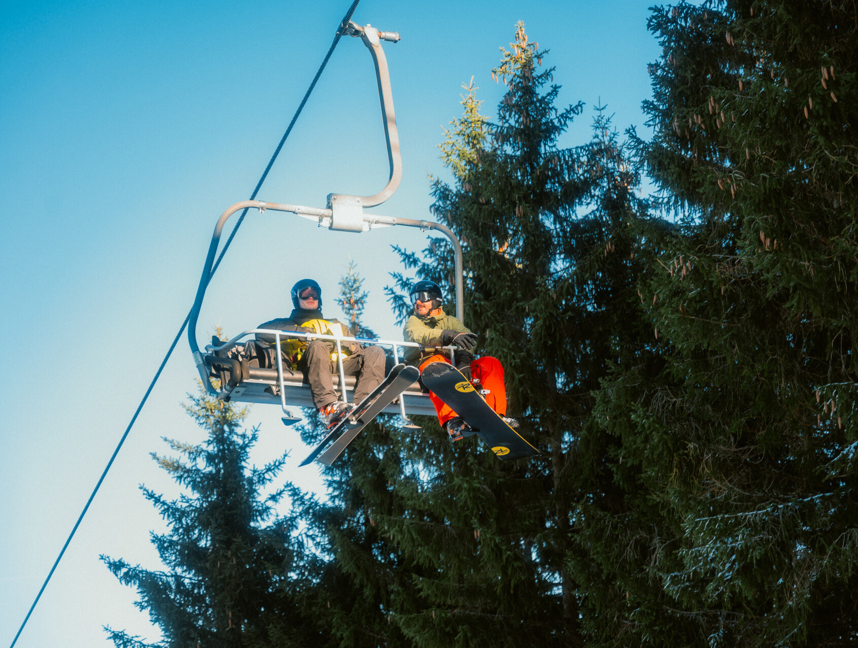 Zwei Snowboarder fahren in Winterberg mit dem Sessellift an Nadelbäumen vorbei.  