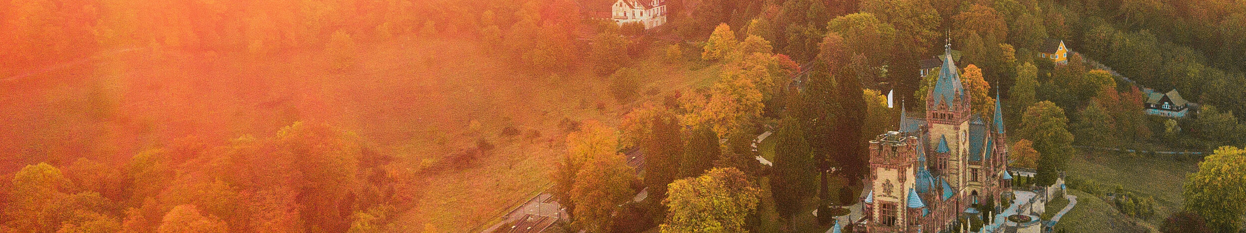 Blick auf das in rötliches Morgenlicht getauchte Schloss Drachenburg bei Königswinter, umgeben von bewaldeten Hügeln im Naturpark Siebengebirge 