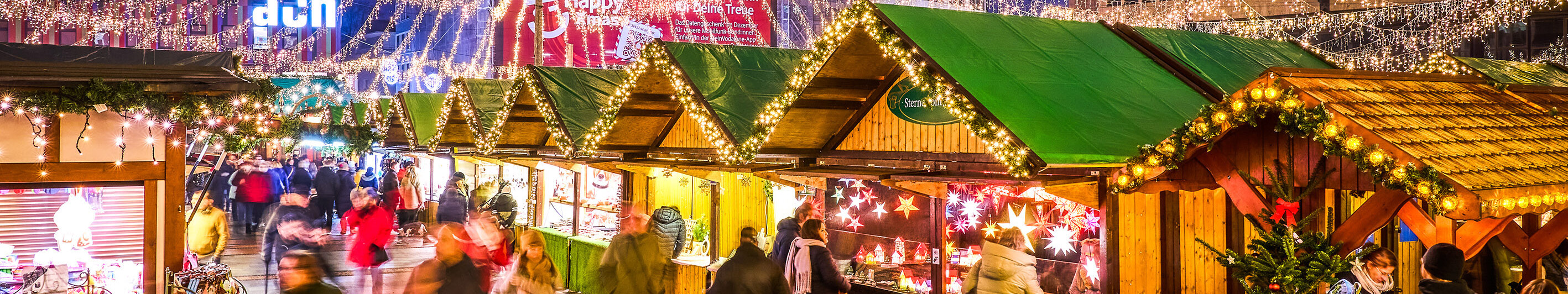 Festlich beleuchteter Internationaler Weihnachtsmarkt in Essen am Abend. Über den Holzhütten sind Lichterketten gespannt, die sich strahlenförmig von einem zentralen Punkt mit einem leuchtenden Stern aus nach außen verteilen und den Markt in ein warmes, gemütliches Licht tauchen. Besucher schlendern durch die Gänge zwischen den Buden, die verschiedene Produkte anbieten. 